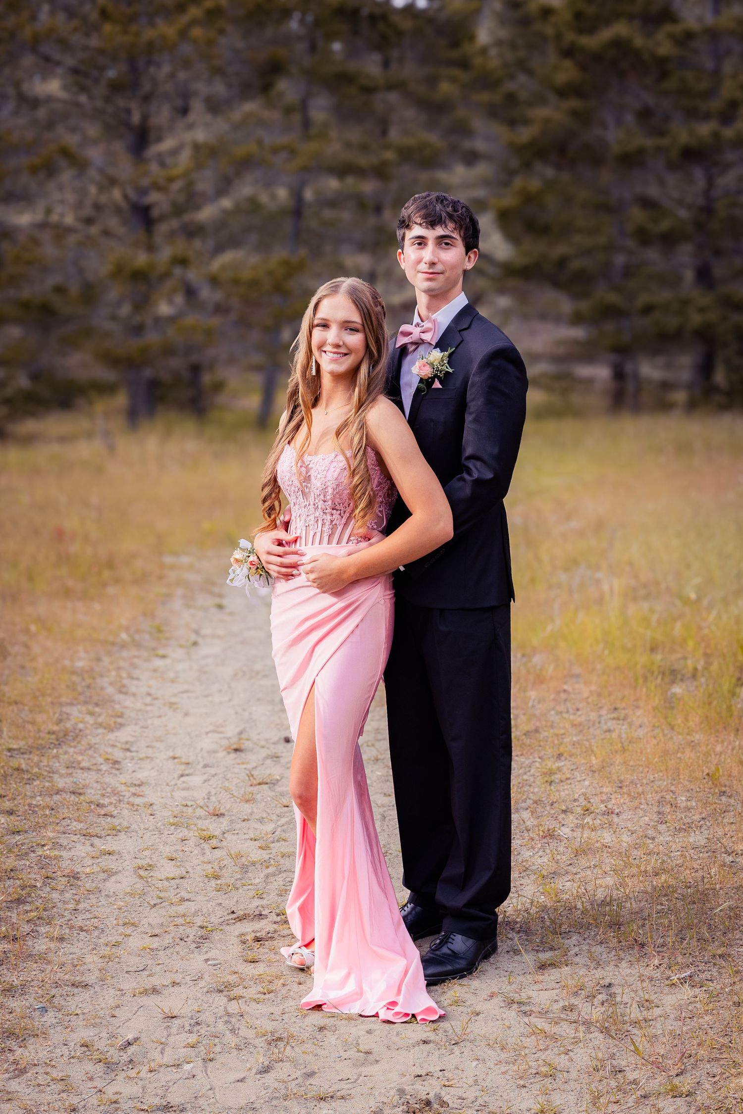 A teenage couple poses in formal prom attire outdoors, she in a pink gown and he in a black suit with bow tie.