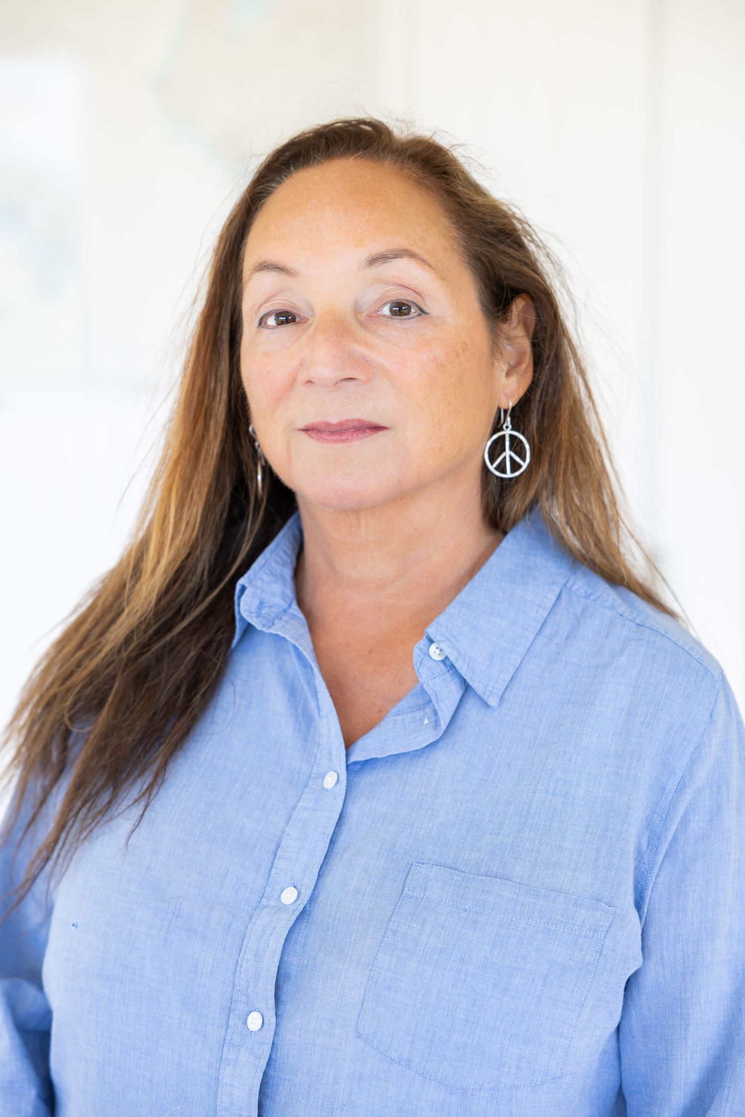 Middle-aged woman with long brown hair wearing a blue shirt and peace sign earrings, smiling softly indoors.