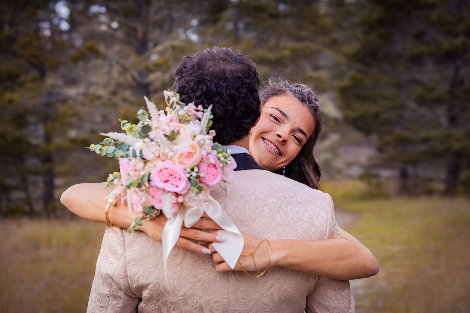 Two women share a joyful hug outdoors, one holding a pink floral bouquet, suggesting a wedding celebration.