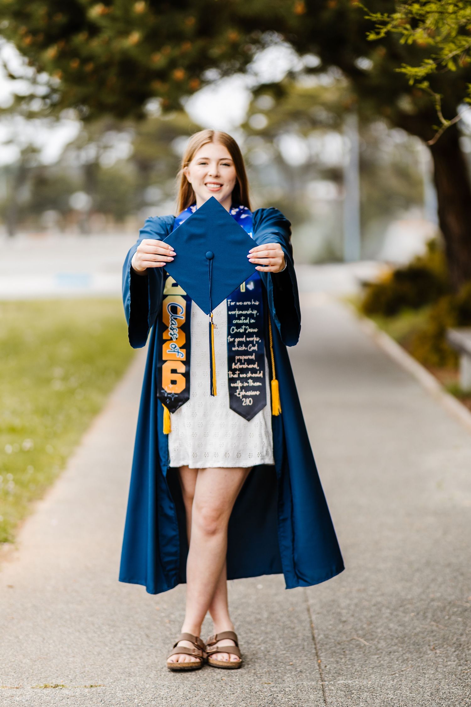 Young woman in blue graduation gown holding decorated cap outdoors on a sunny day.