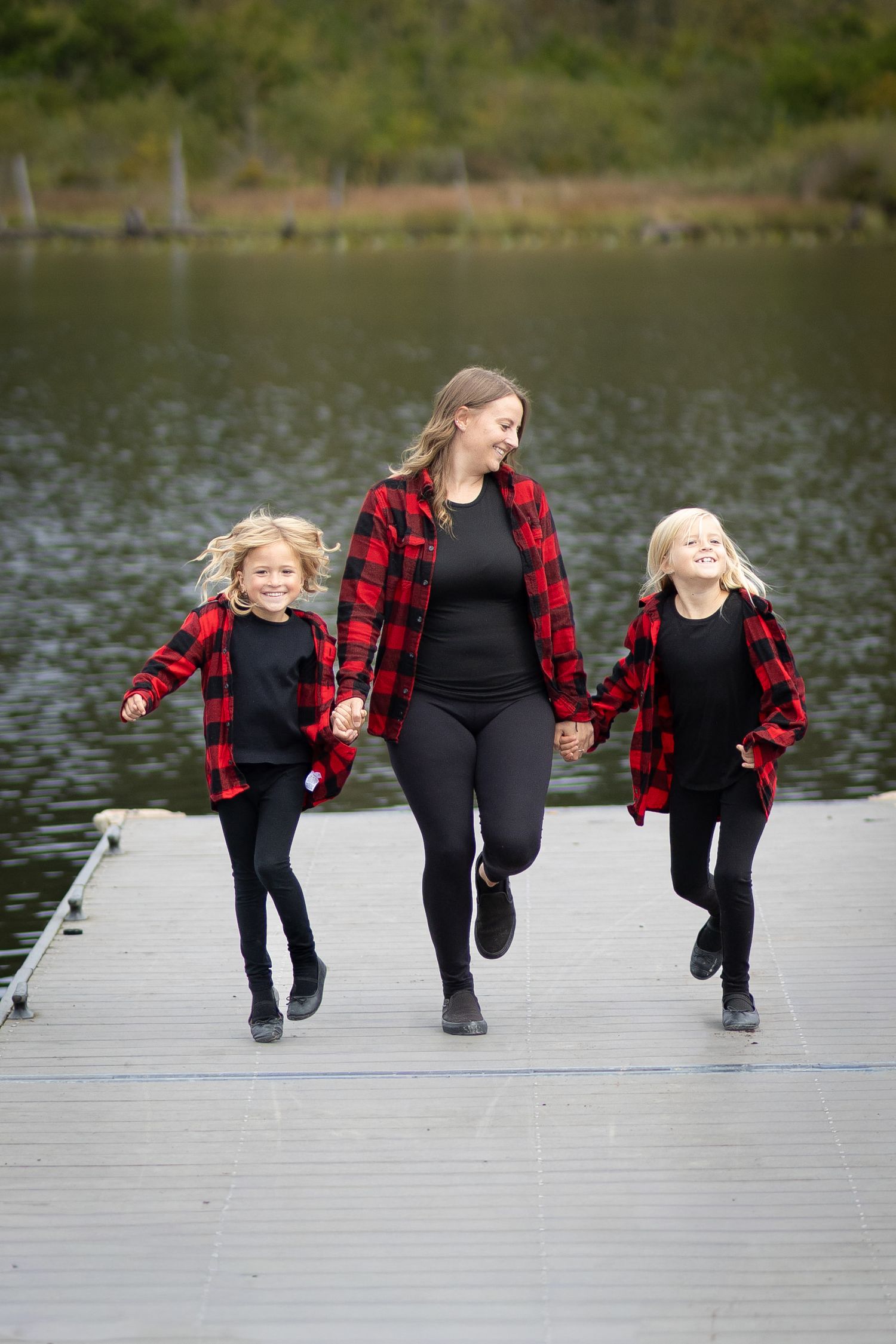 Mother and two daughters in matching red plaid shirts run hand-in-hand on a dock by a lake.