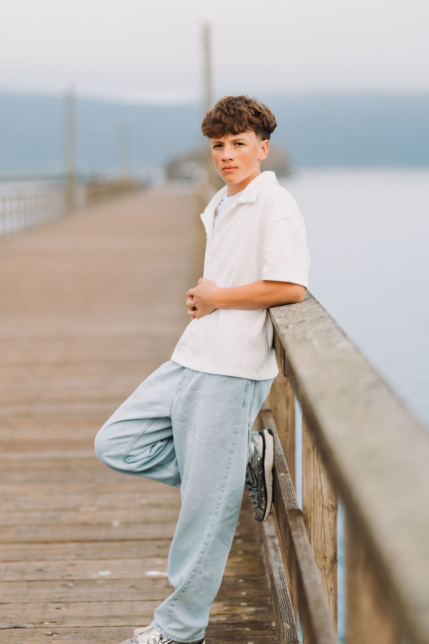 Teen boy in white polo and light blue joggers leaning on wooden pier railing with water in background.