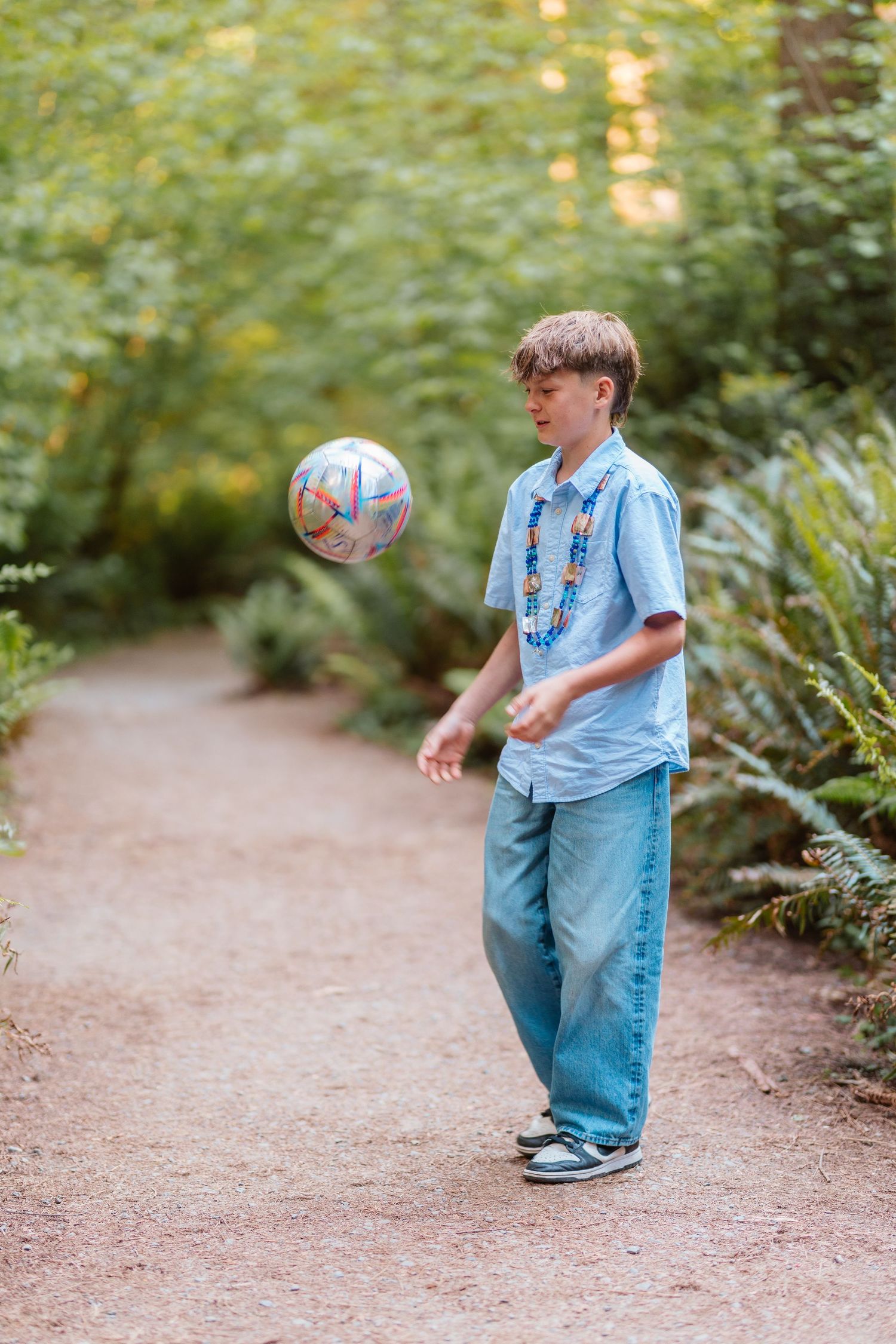 A boy juggling a soccer ball on a forest path, wearing a light blue shirt, beaded necklace, and jeans.