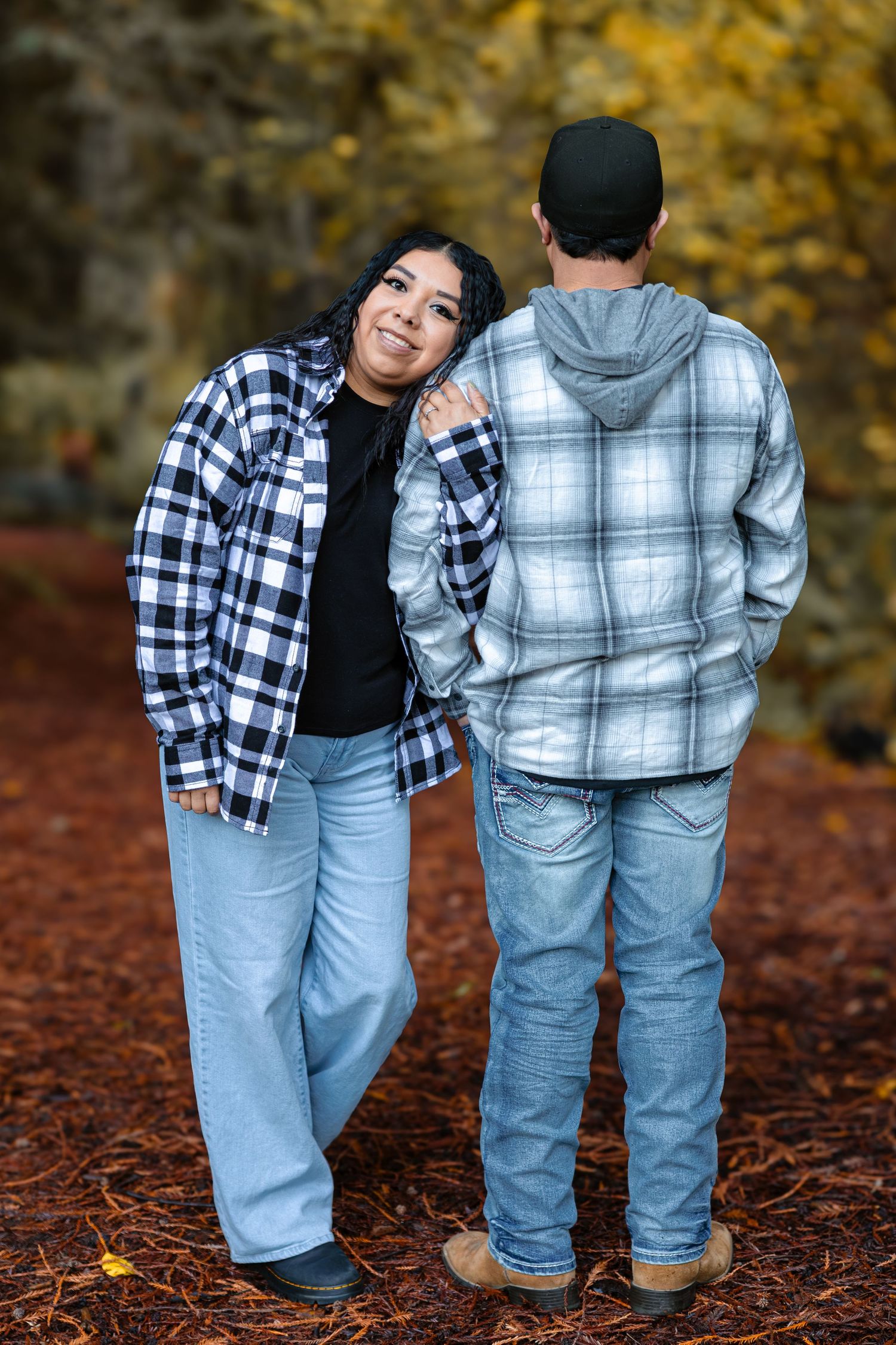 Woman in black and white plaid shirt smiles at camera while leaning on mans shoulder in autumn redwood forest.