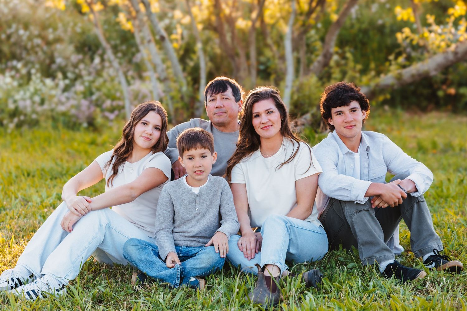 Family of five sitting on green grass outdoors, smiling together with golden sunset trees in the background.