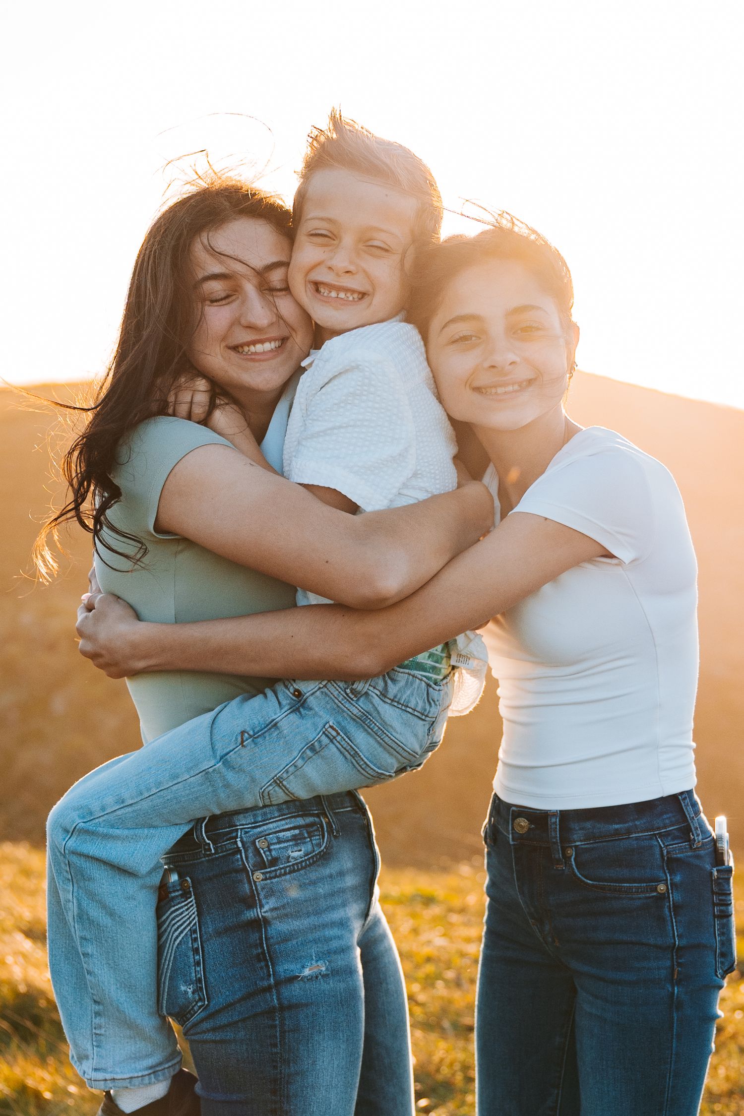 Three siblings hugging and laughing together in golden sunlight on a grassy hillside.