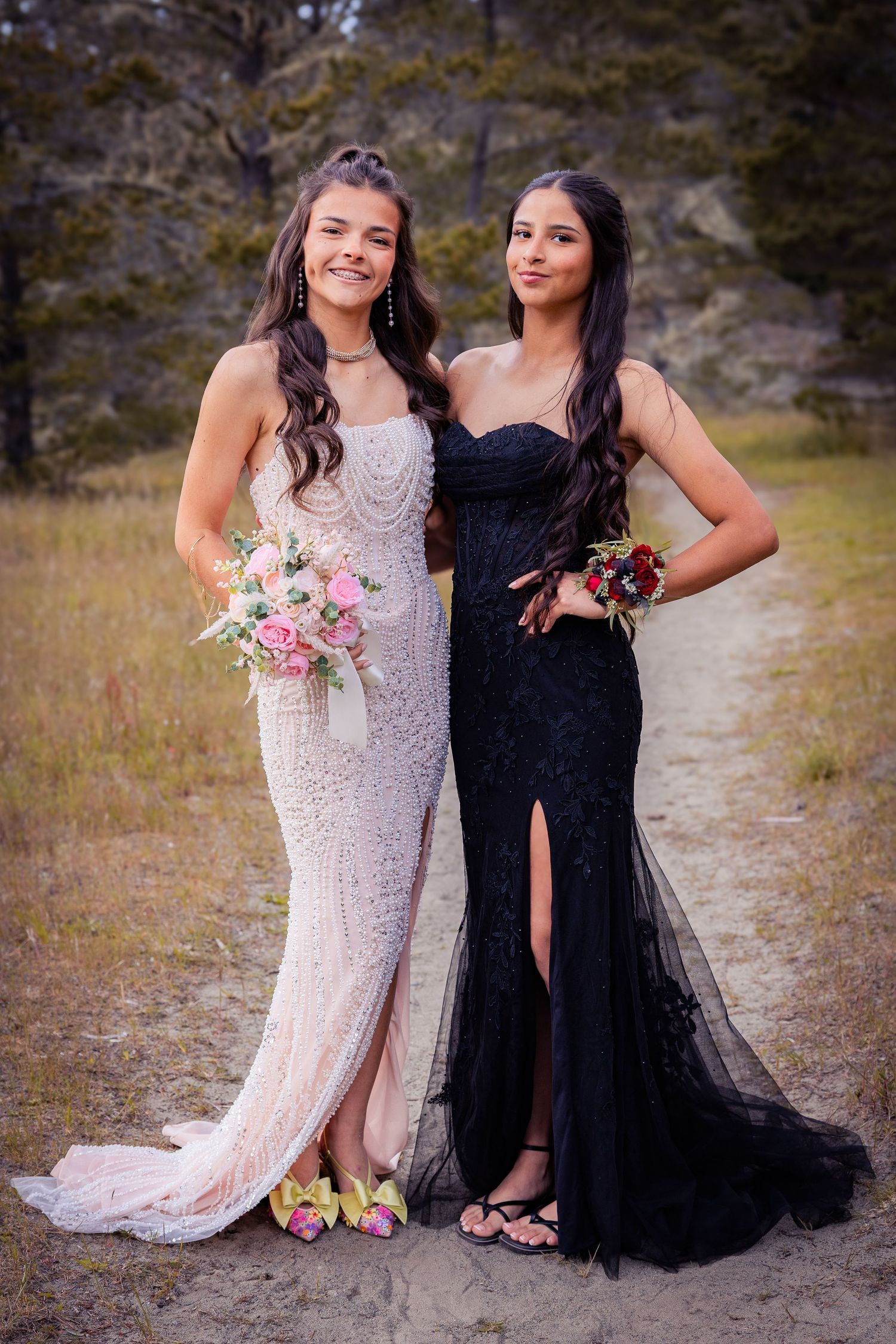 Two young women in elegant formal gowns, one white and one black, holding bouquets outdoors.