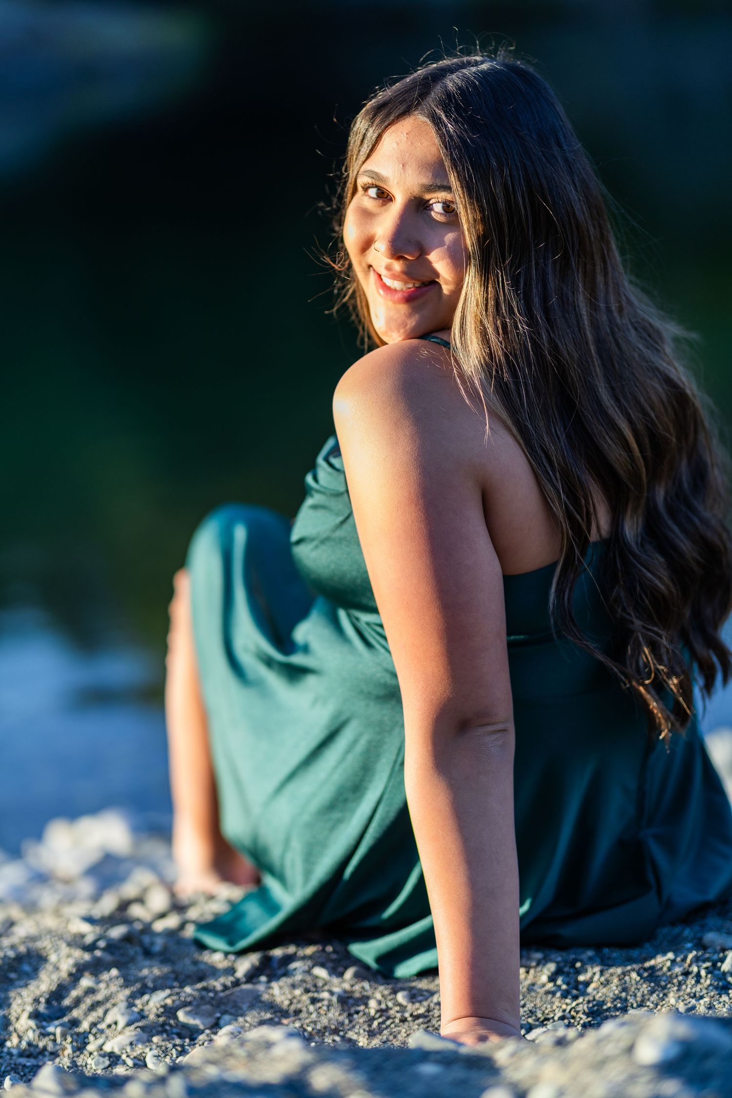 Smiling young woman in green dress sitting on rocks by a reflective lake at sunset.