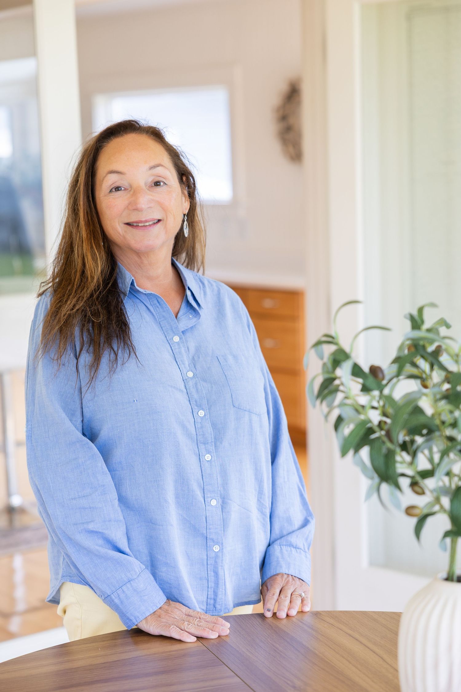 Smiling middle-aged woman with long brown hair wearing a blue shirt, standing at a wooden table in a bright home interior.