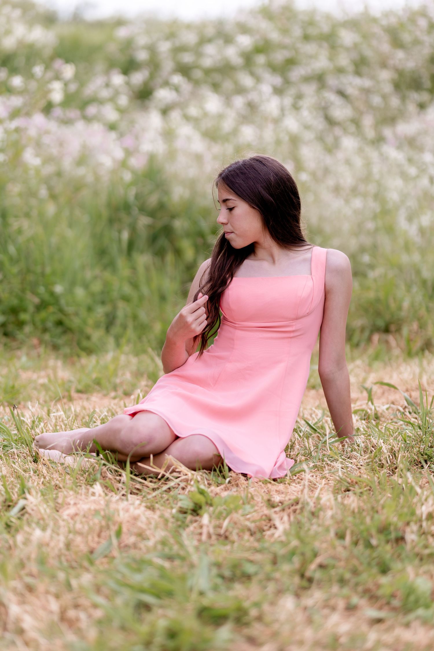 Young woman in pink sundress sitting barefoot in a grassy meadow with wildflowers, looking down thoughtfully.