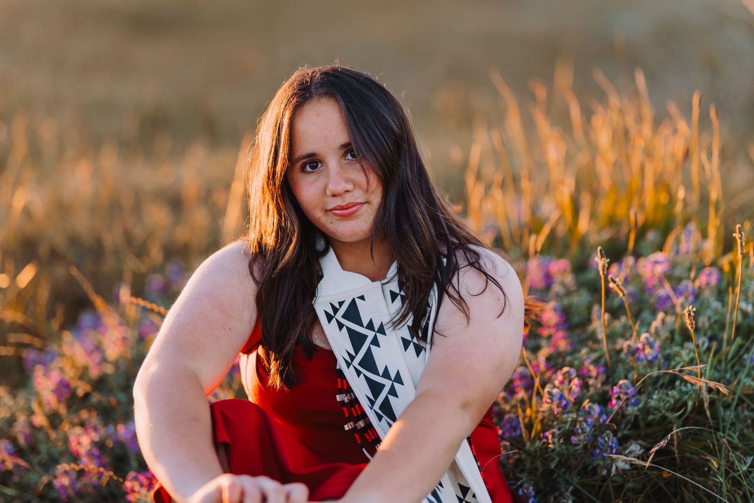 Young woman with dark hair sits among purple wildflowers in a golden field at sunset, wearing a red and white dress.