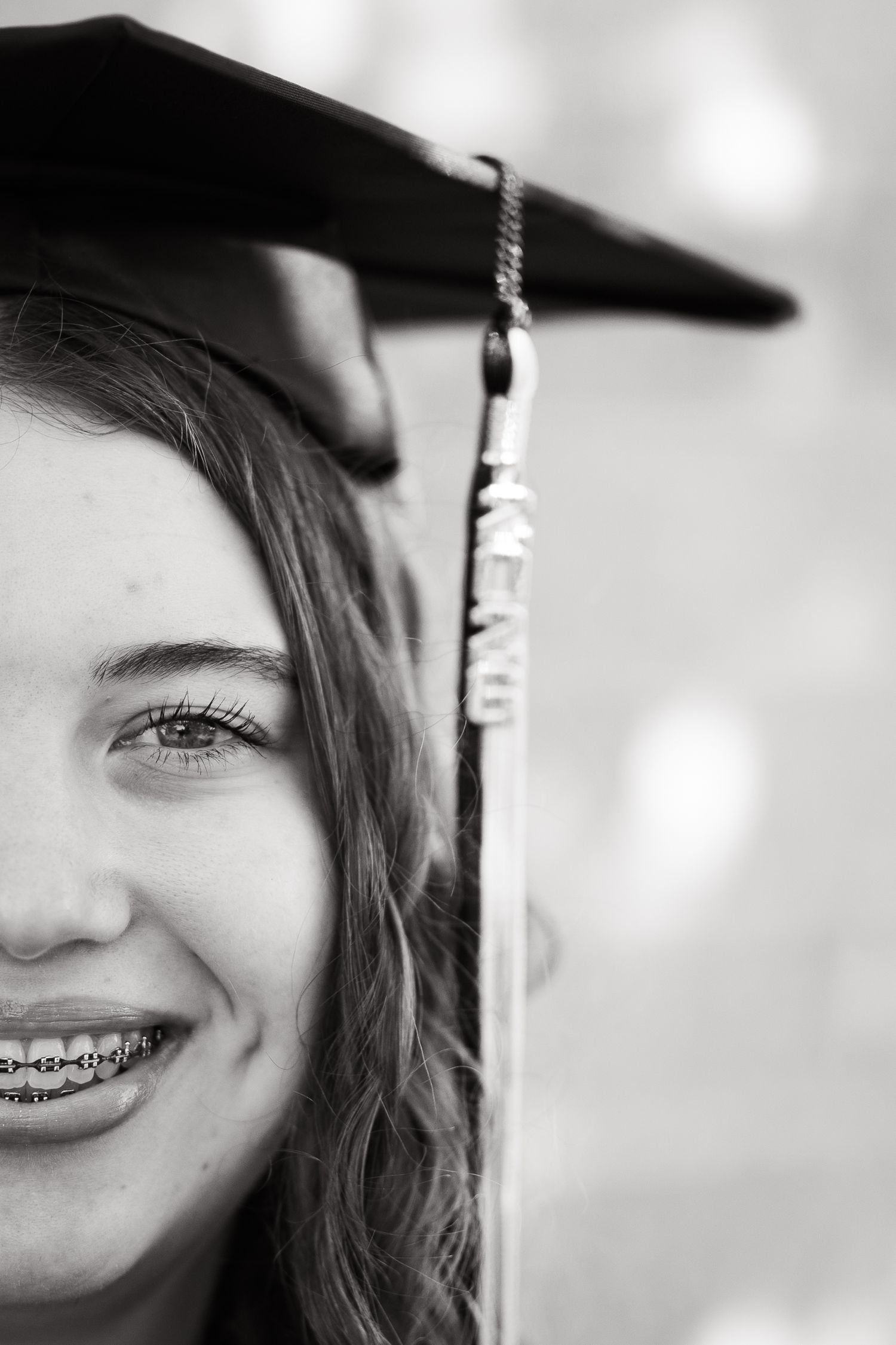 Smiling young woman wearing graduation cap and braces in black and white portrait photo.