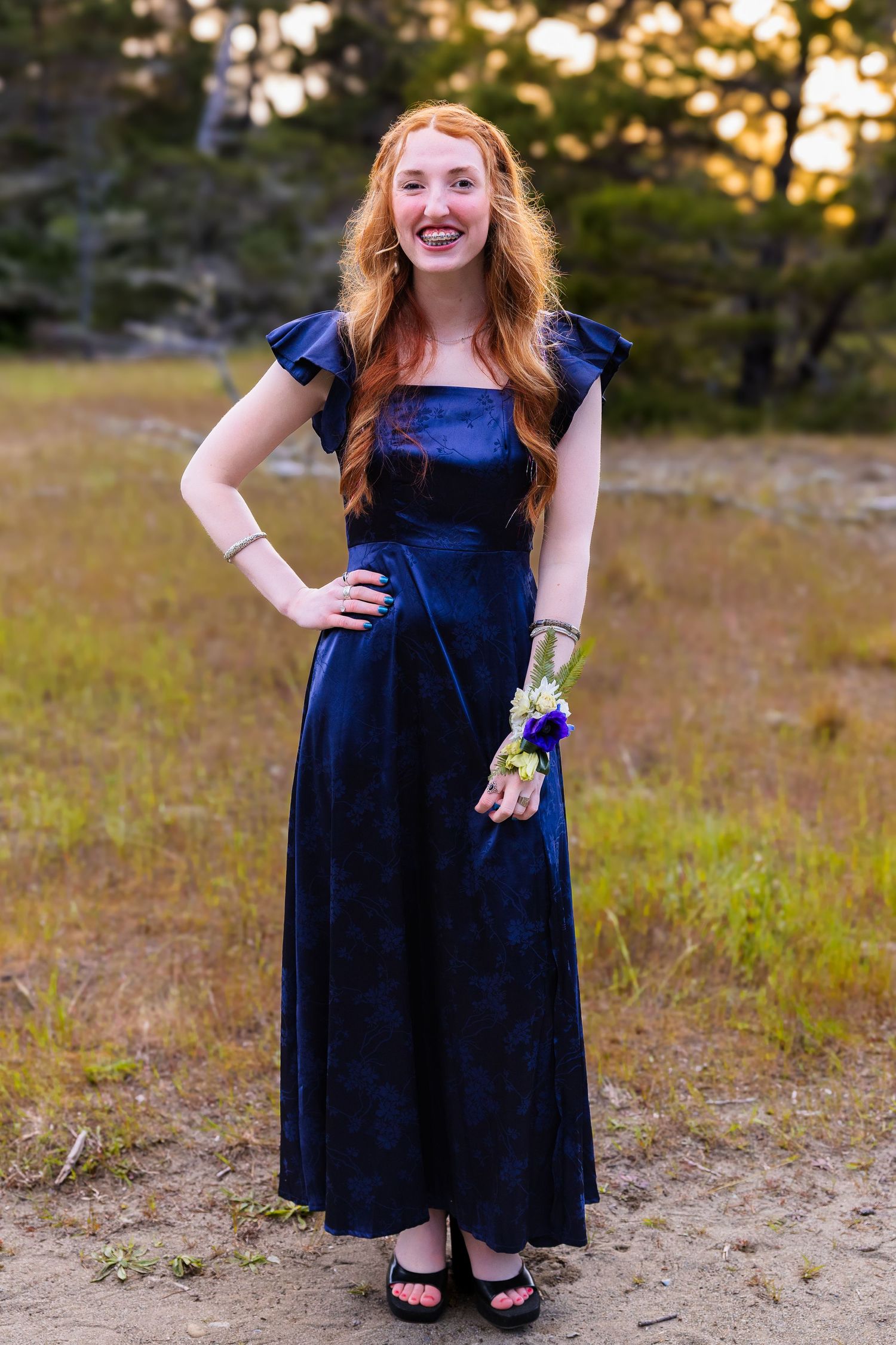 Young woman in navy blue velvet gown with floral wrist corsage smiling outdoors at prom.