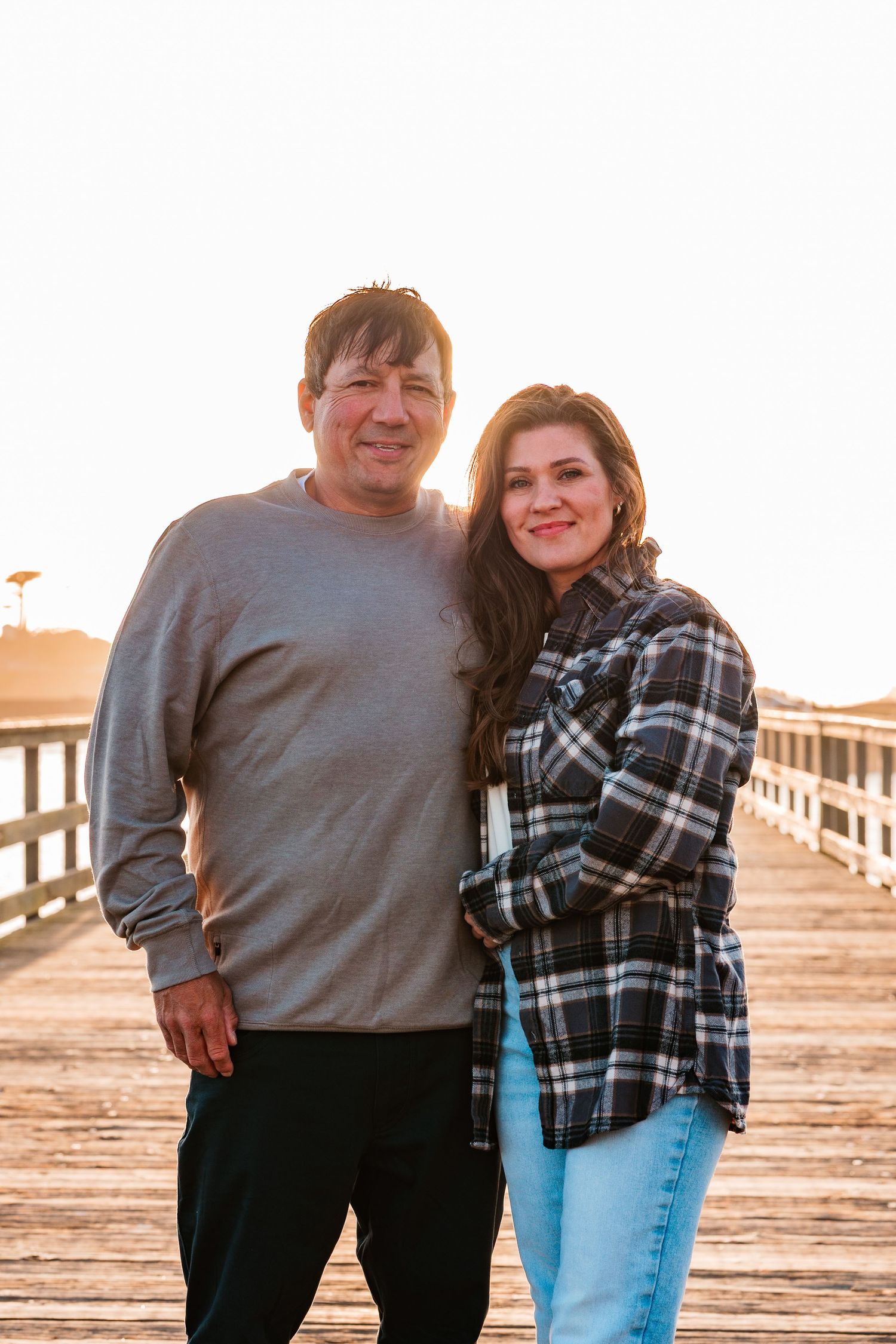 Couple smiling together on a wooden pier during golden hour sunset at the beach.