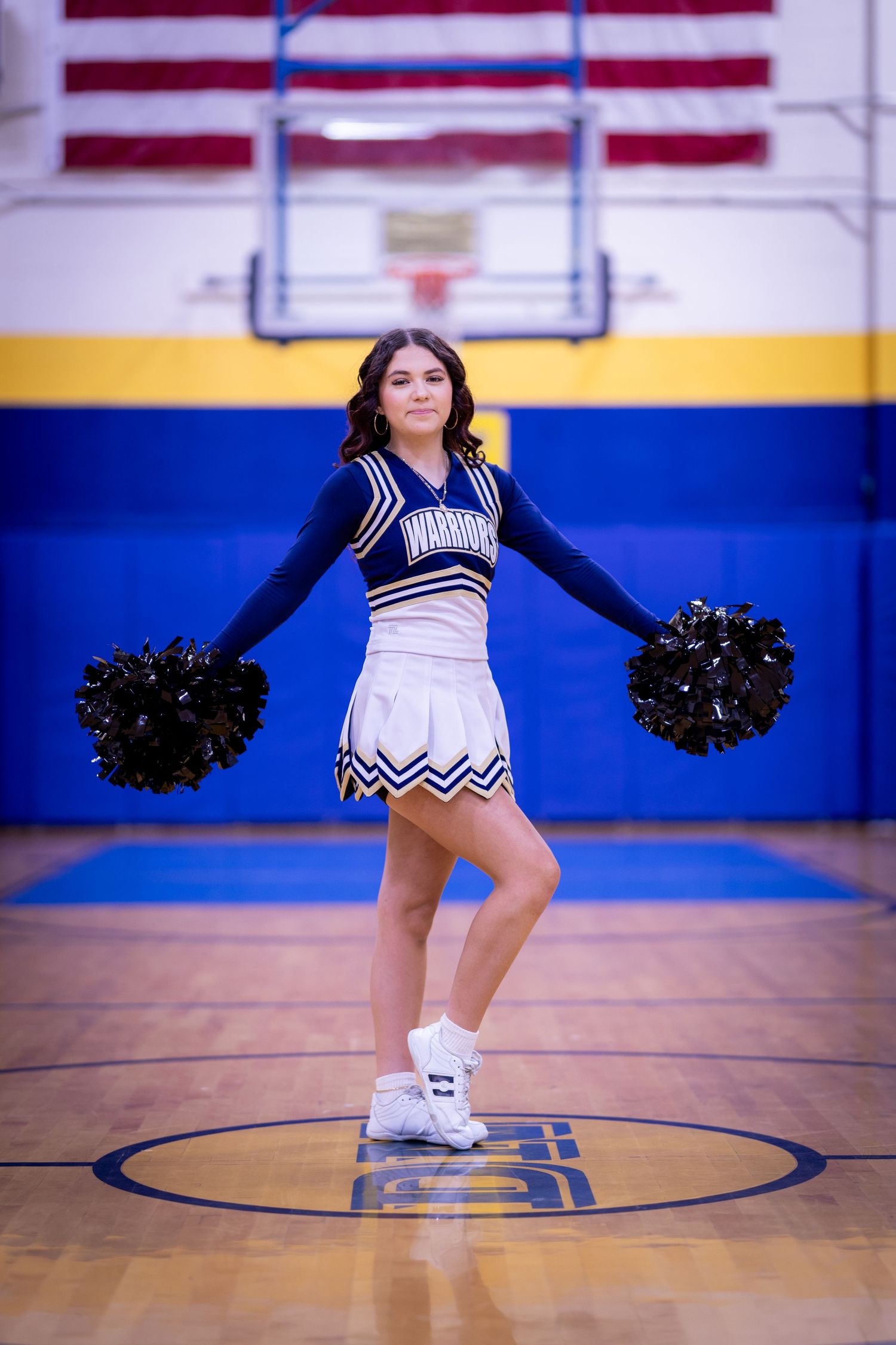 Warriors cheerleader in navy uniform poses with black pom-poms in gym.