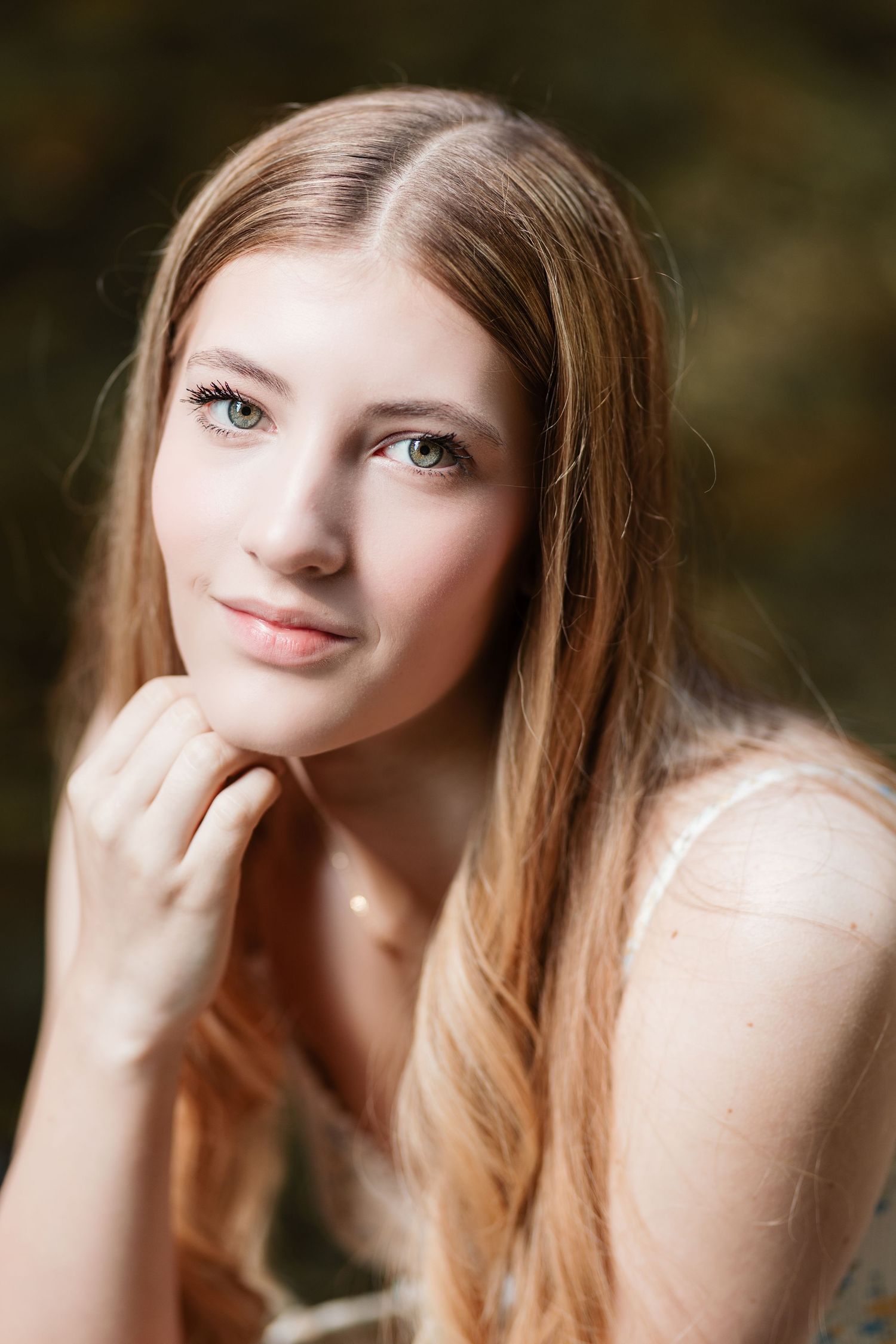 Young woman with long auburn hair resting chin on hand with green eyes in natural outdoor setting.