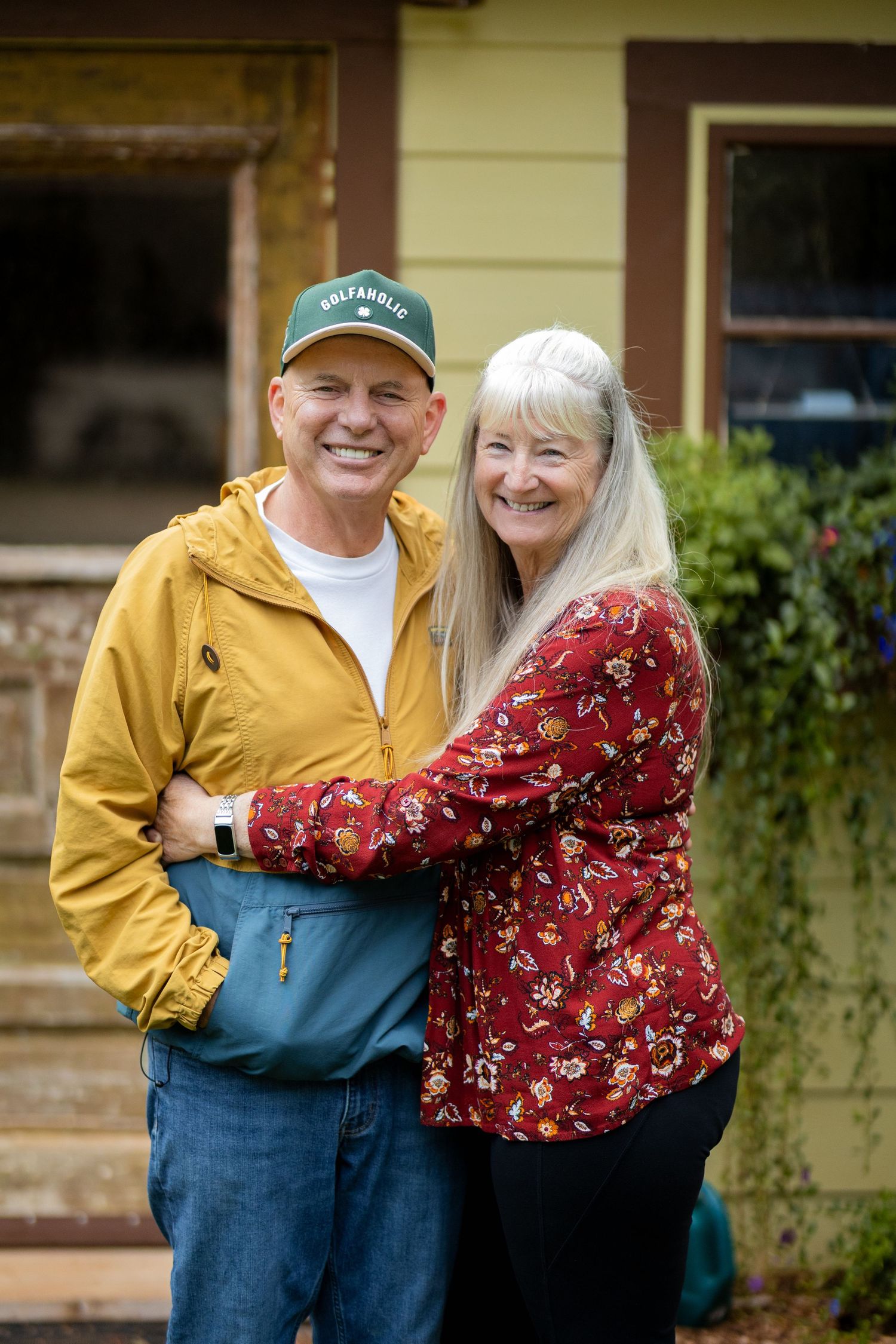 A happy older couple embracing and smiling in front of a house with green siding and plants.