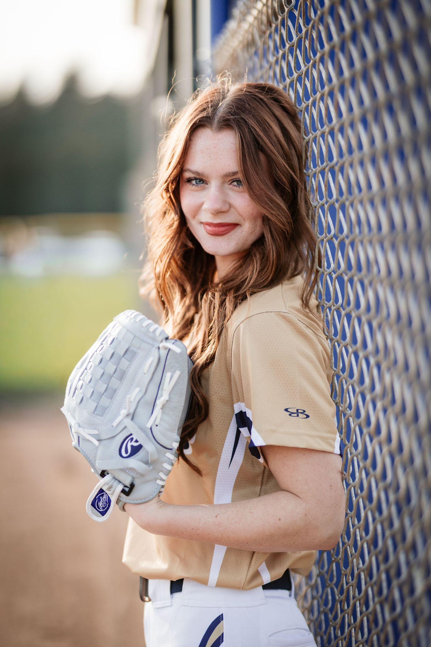 Female softball player in gold uniform leaning against chain-link fence holding a white fielding glove.
