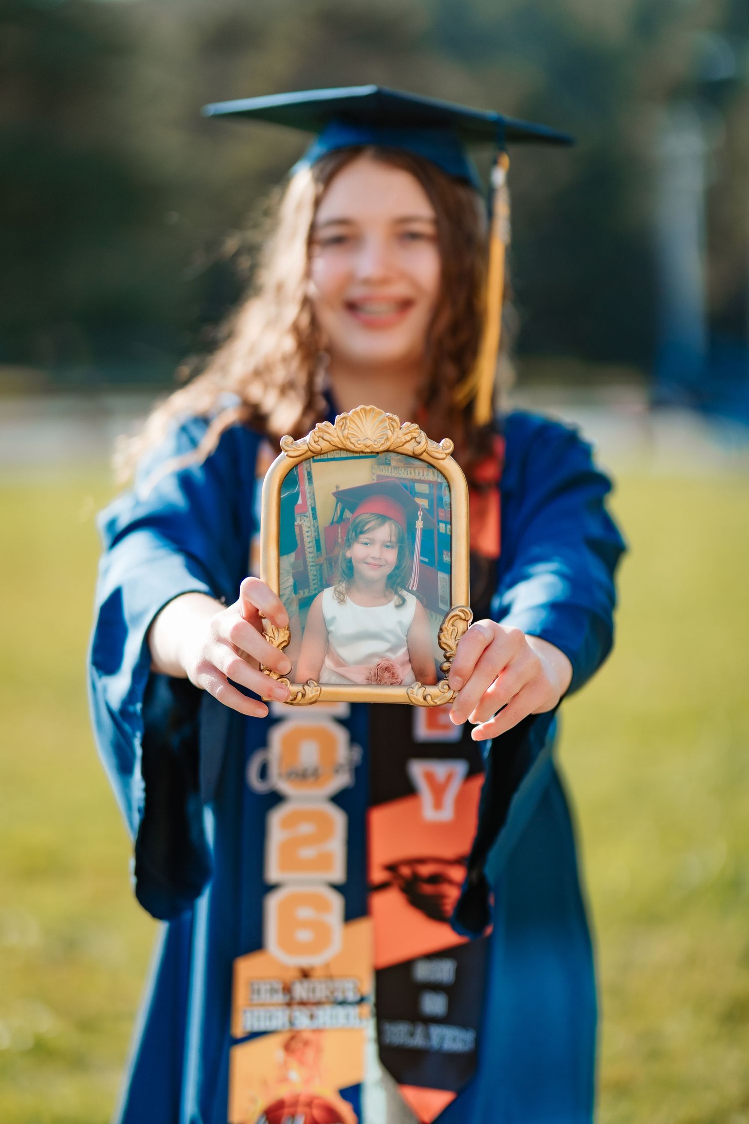 Graduate in blue cap and gown holds framed childhood photo, smiling outdoors during graduation celebration.