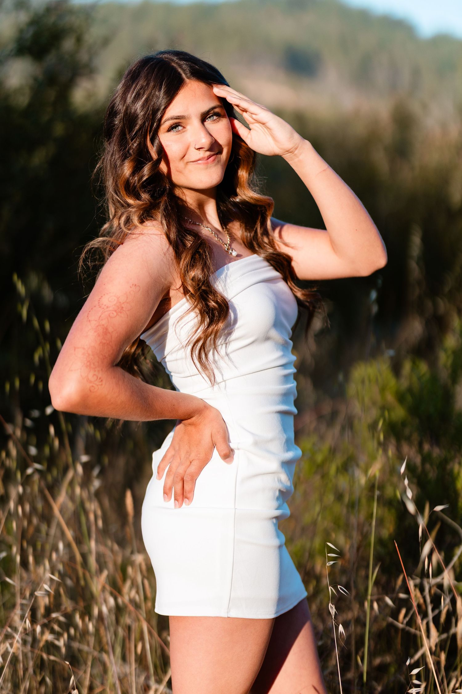 Young woman in white mini dress poses in golden field with mountains, shielding eyes from sunlight.