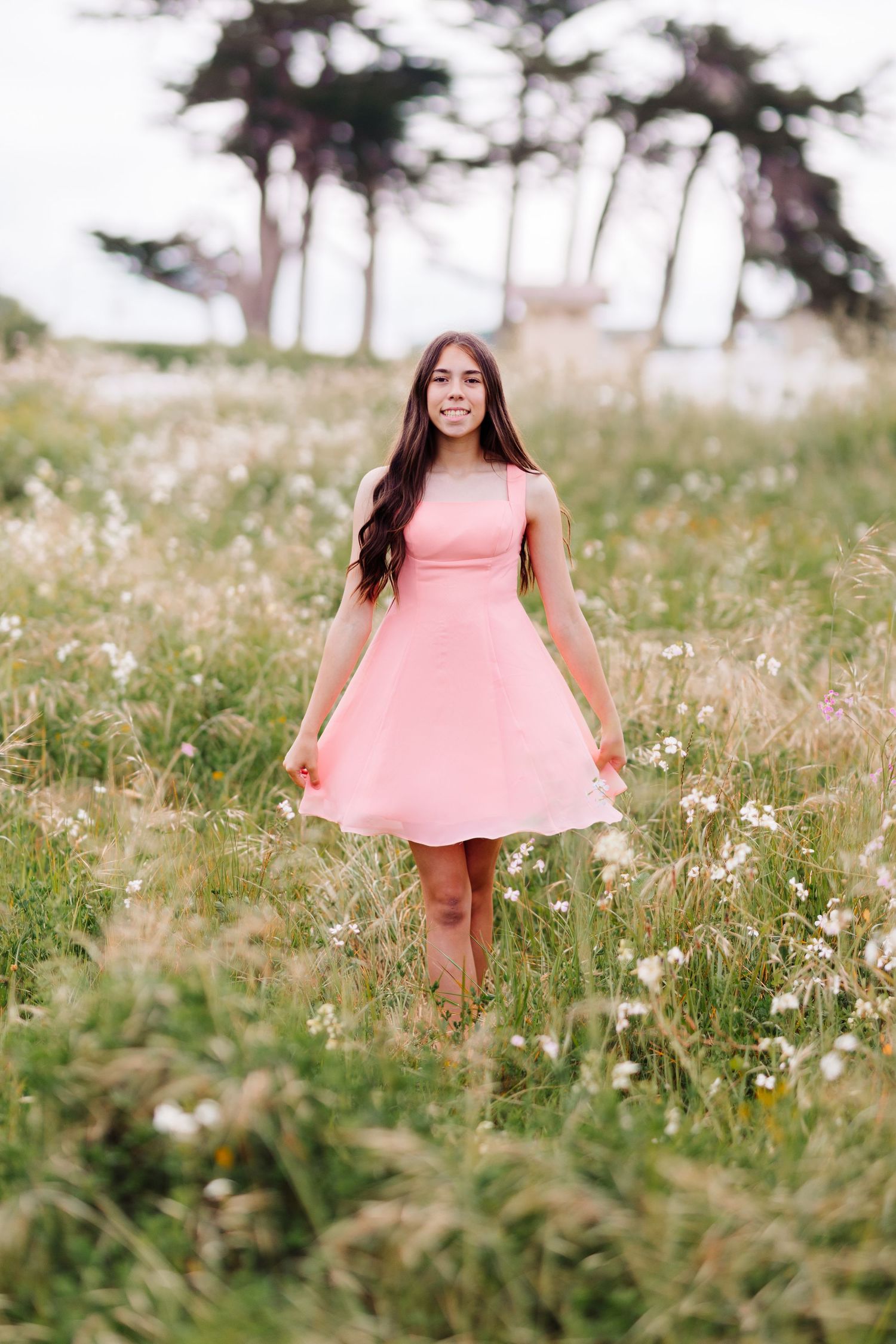 Young woman in a flowing pink dress walking through a wildflower meadow with trees in the background.