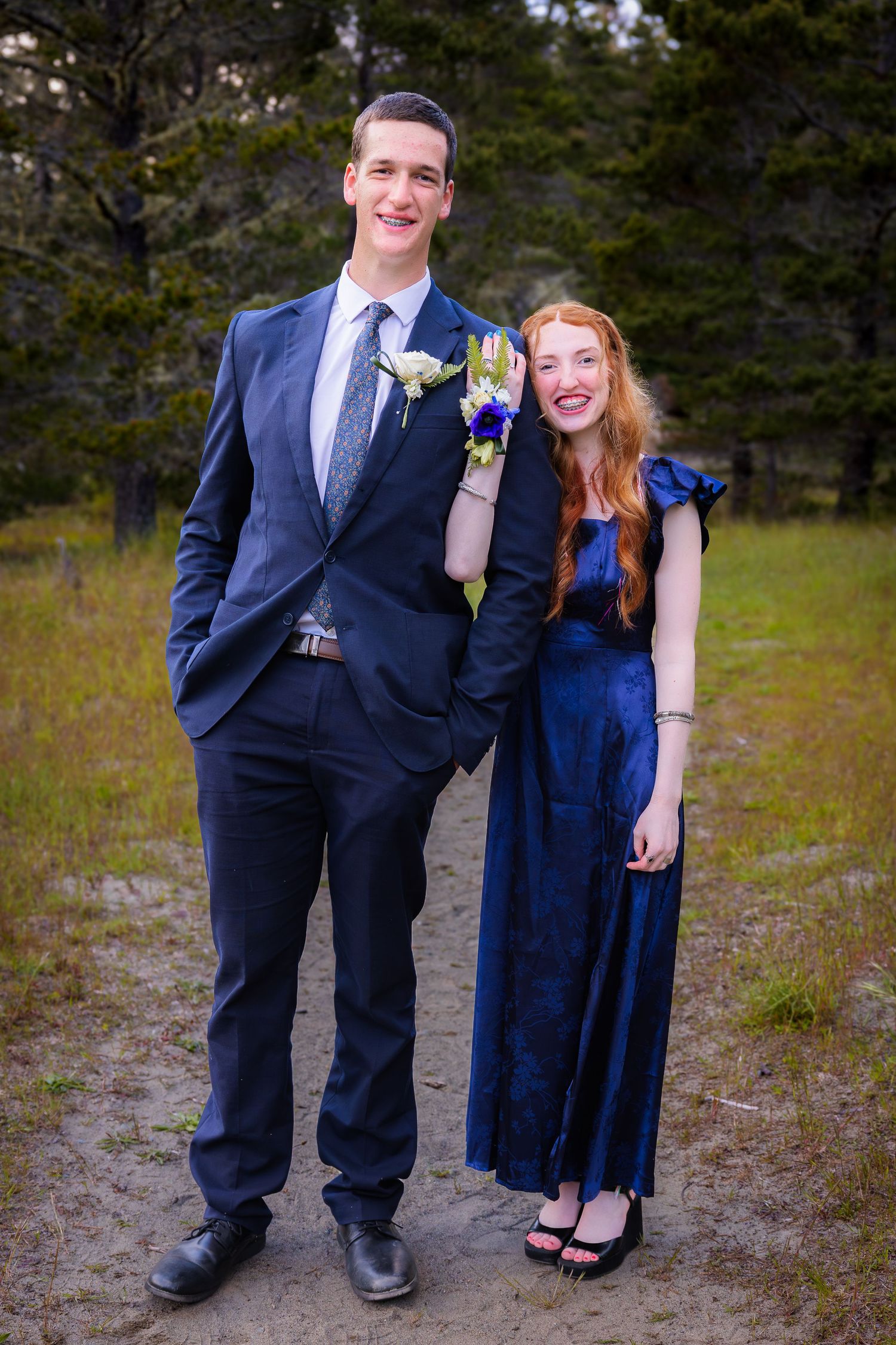 A tall young man in a navy suit and a young woman in a blue gown pose together outdoors for prom photos.