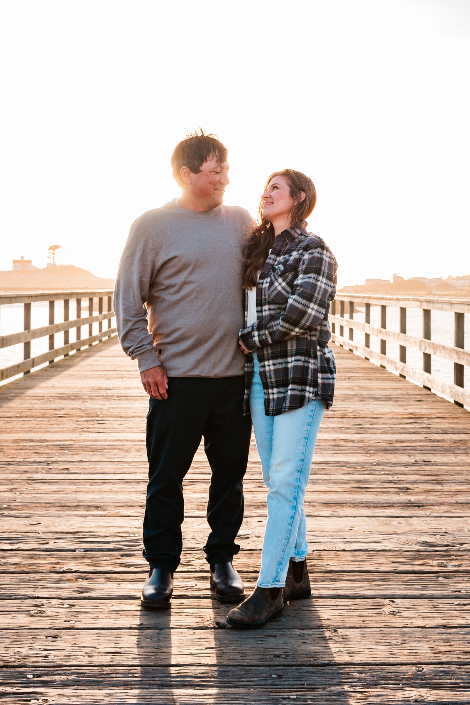 Couple sharing a romantic moment on a sunlit wooden pier, woman in plaid shirt and jeans smiling up at man in gray sweater.