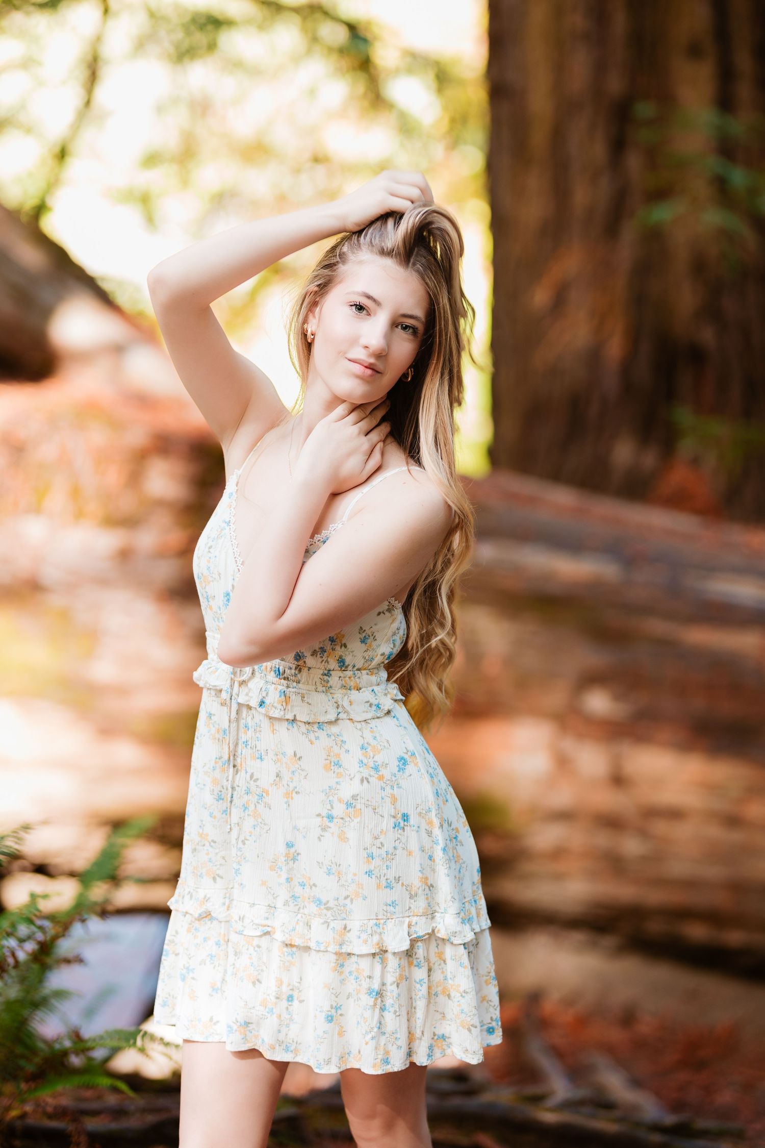 Young woman in a floral sundress posing with hand in hair among tall redwood trees outdoors.