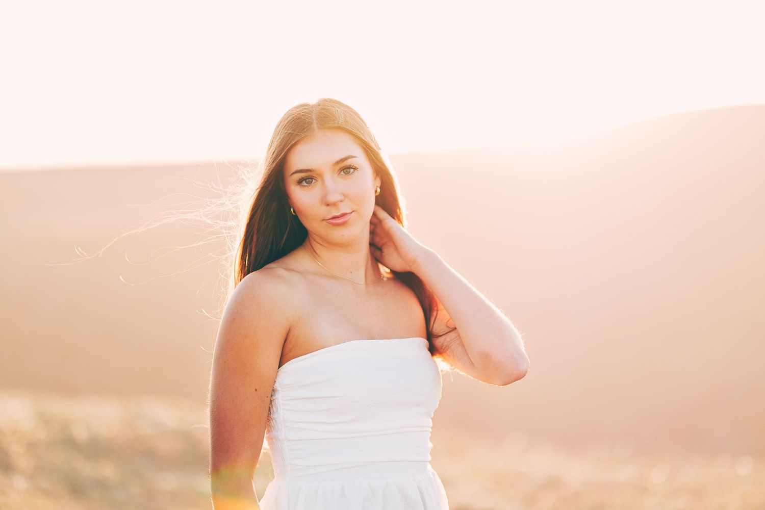 Young woman in a white strapless dress posing in a sunlit field, gently touching her neck.