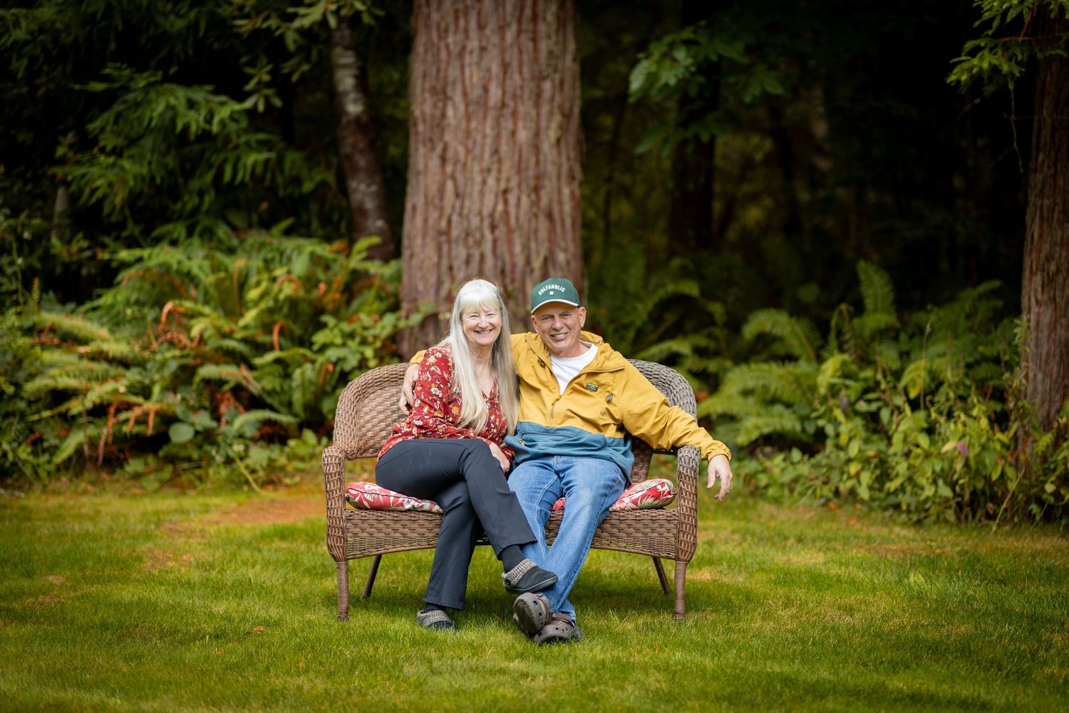 A cheerful older couple sitting together on wicker furniture in a lush green garden setting.