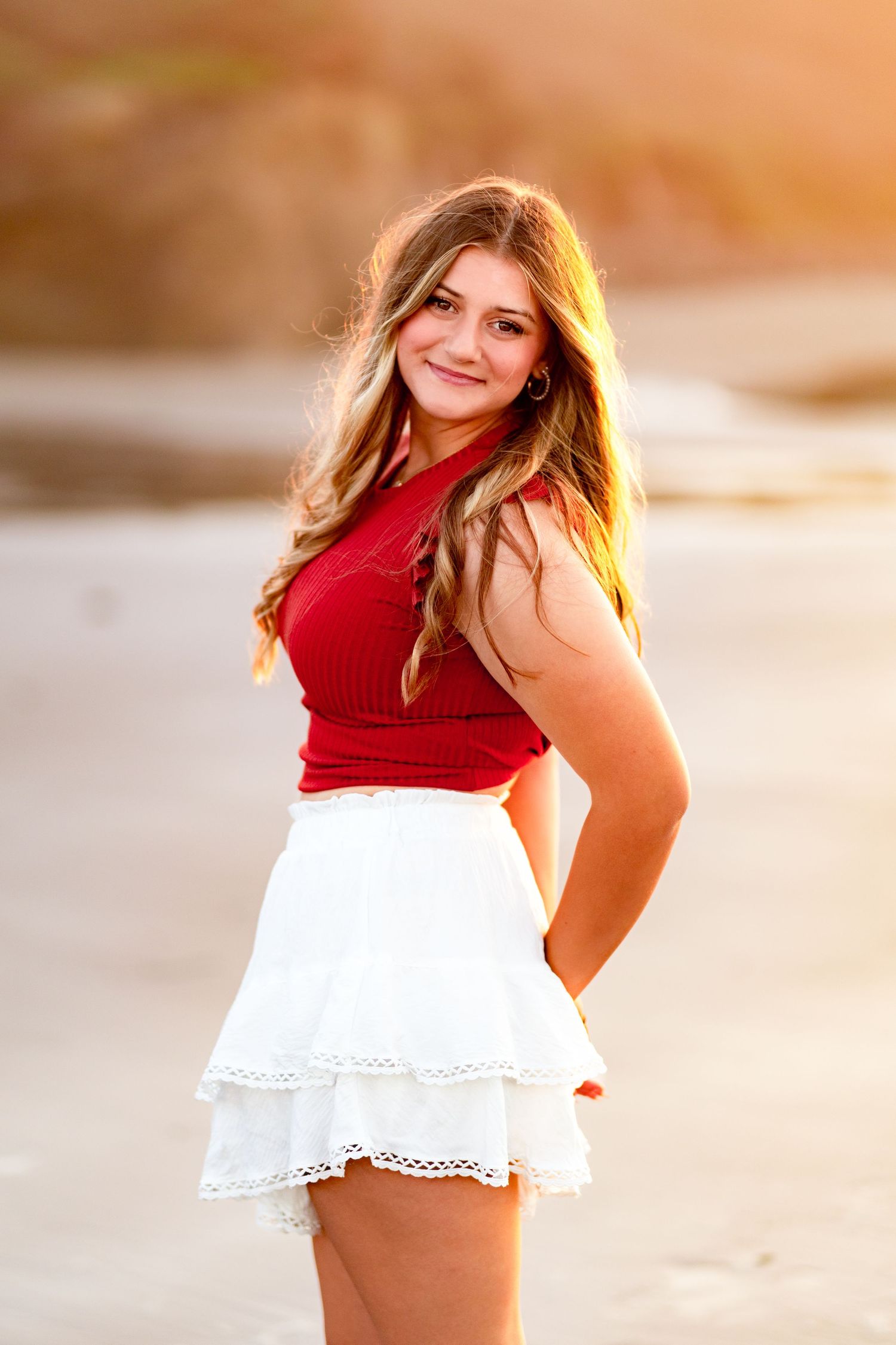 Smiling teen girl in red top and white ruffle skirt posing on beach during warm golden sunset.