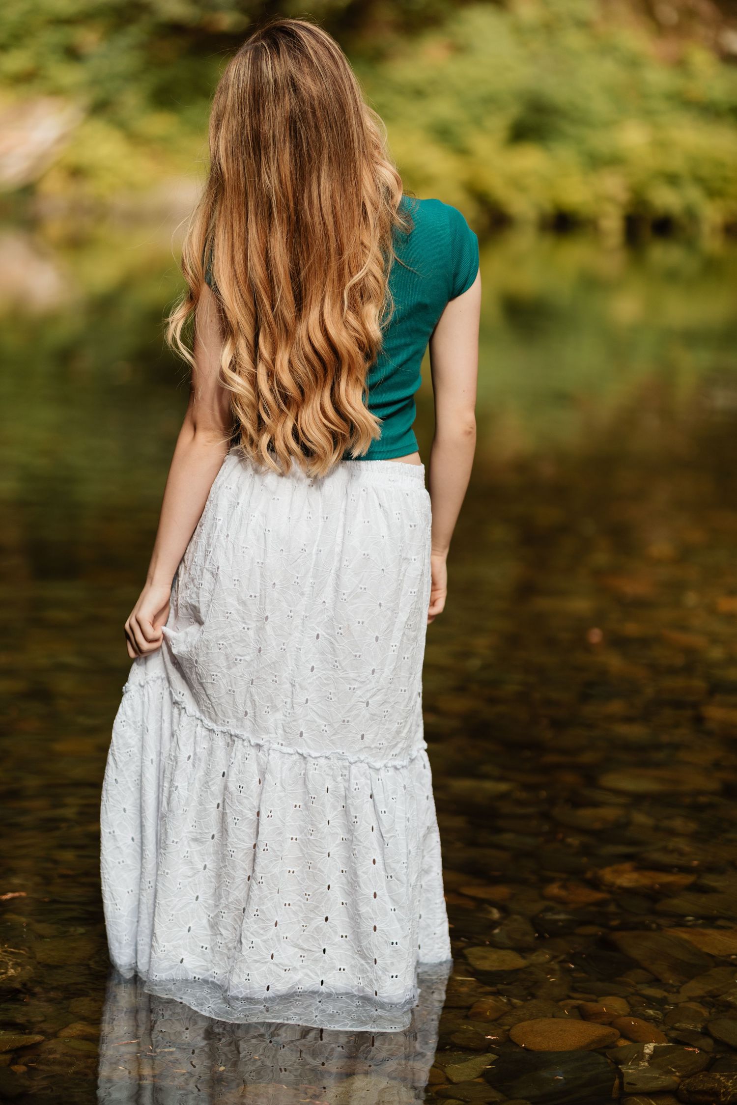 Young woman with long wavy hair in teal top and white skirt standing in a creek.
