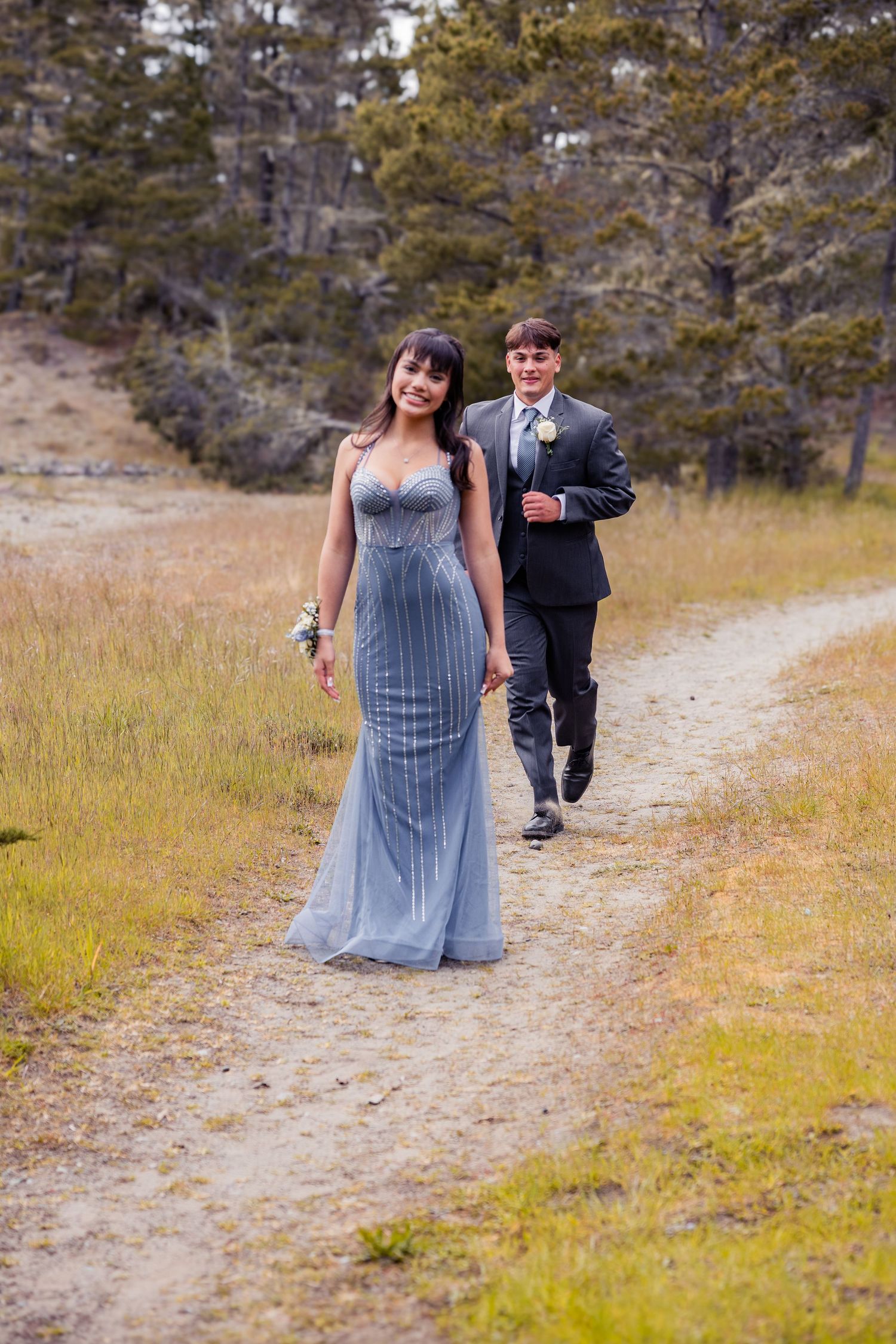 A young couple in prom attire walks a dirt path, she in a blue gown, he in a dark suit with boutonniere.