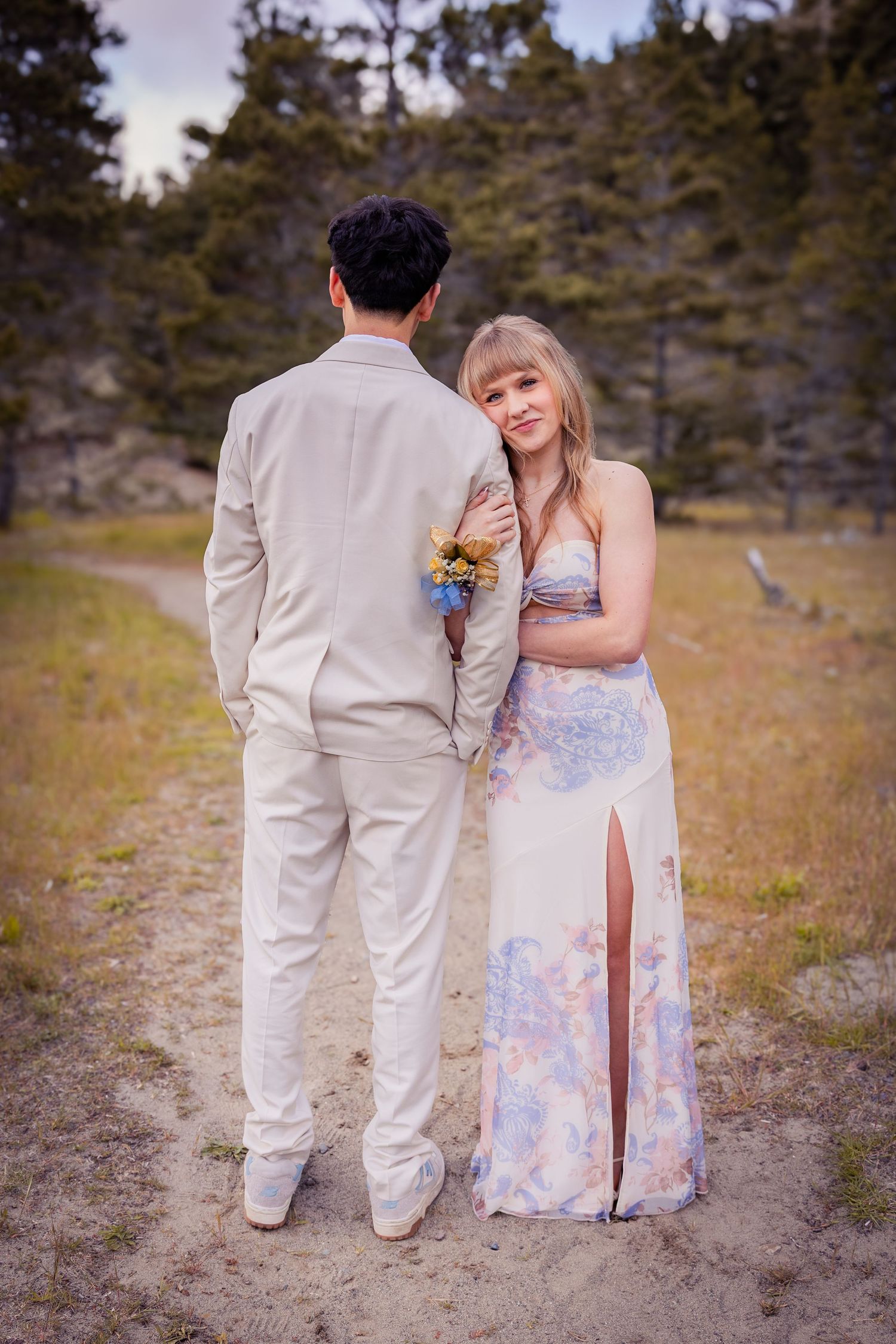 Couple in formal attire standing outdoors, woman in floral dress leaning on man in beige suit, pine trees in background.