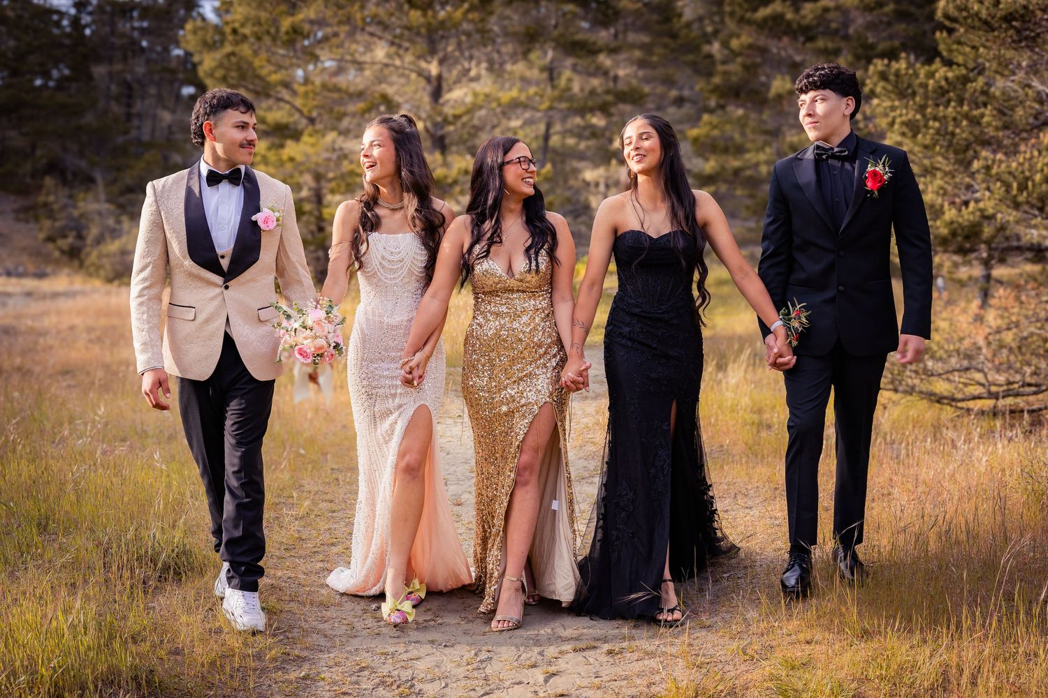 Five teens in formal prom attire walk outdoors, wearing elegant gowns and suits in gold, black, and white tones.
