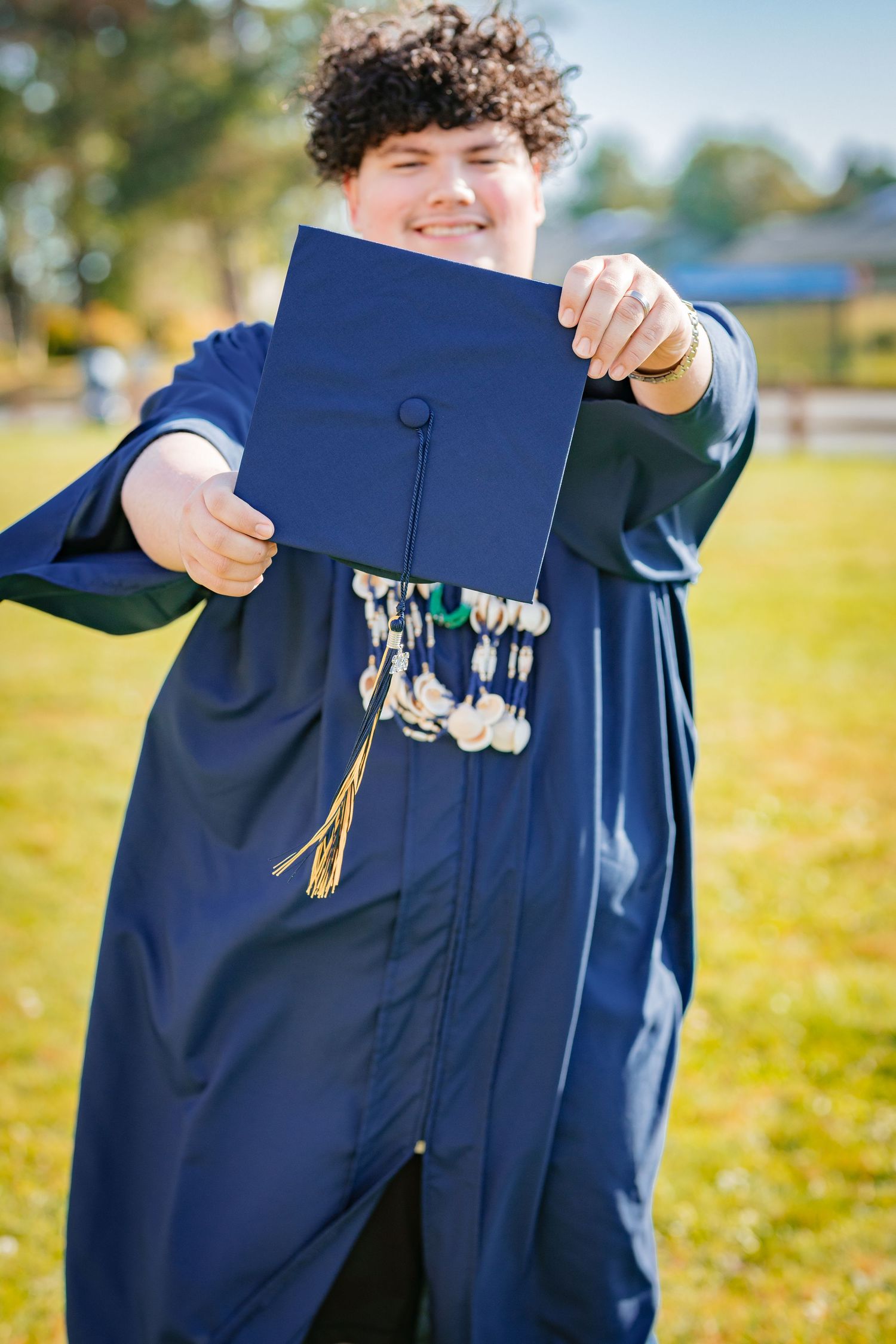 Smiling graduate in navy blue cap and gown holding graduation cap with tassels outdoors on a sunny day.