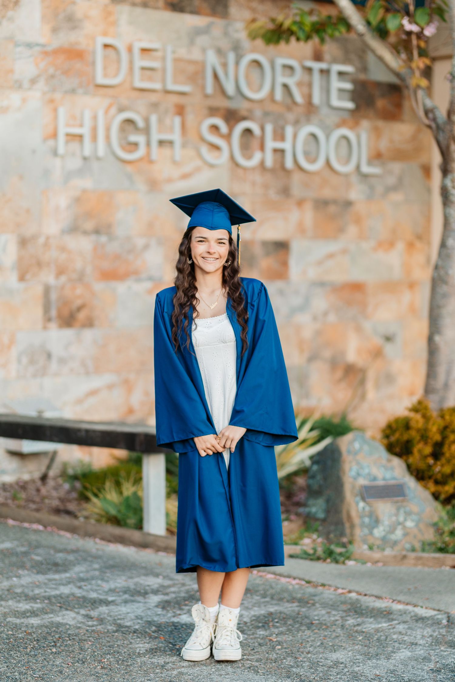 A smiling girl in a blue graduation cap and gown stands in front of Del Norte High School sign.