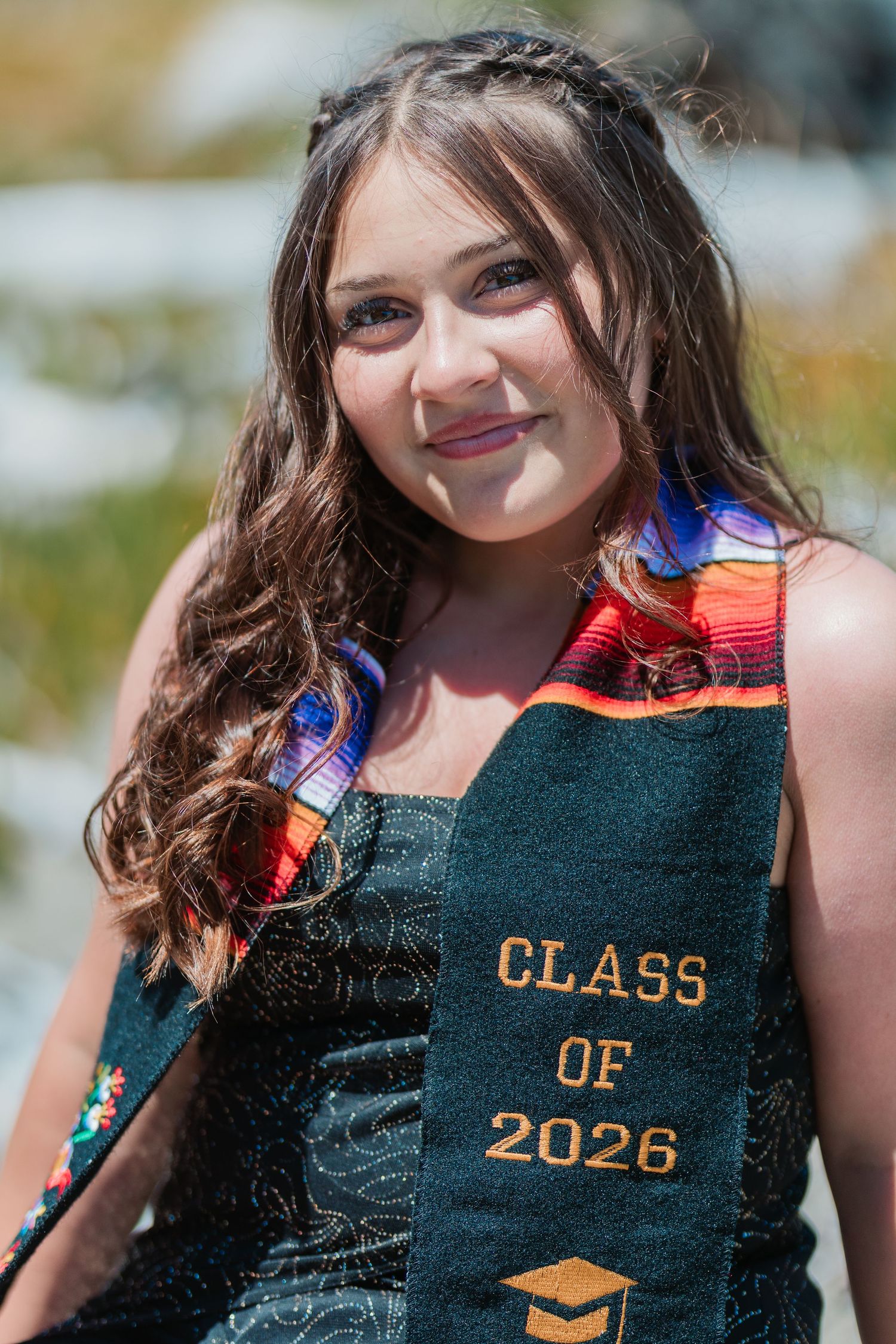 Young woman wearing a Class of 2026 graduation stole and colorful sash, smiling outdoors in bright sunlight.