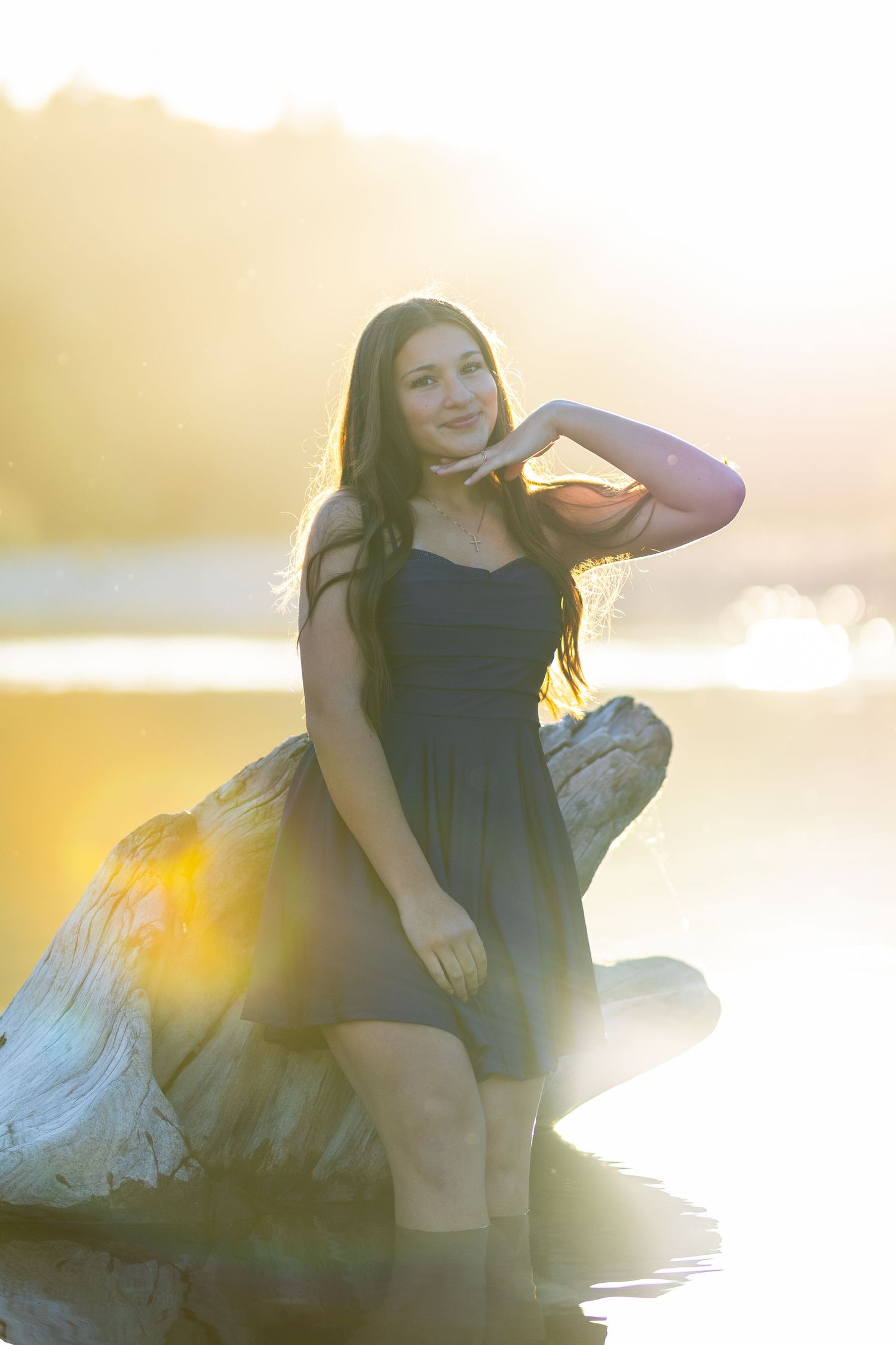 Young woman in black dress sitting on driftwood by a lake at golden hour sunset.