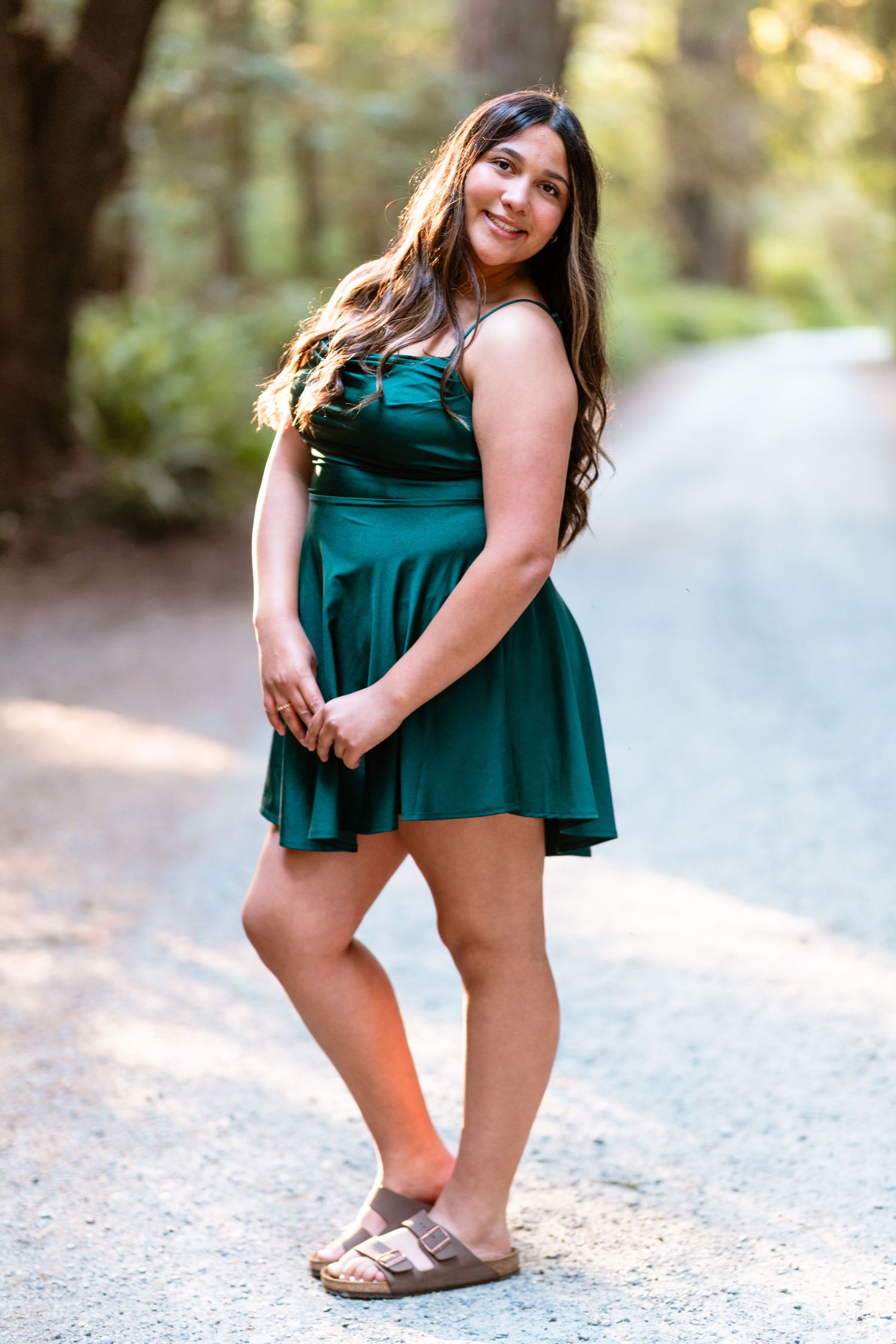 Young woman in green mini dress and sandals smiling on a sunlit forest path.