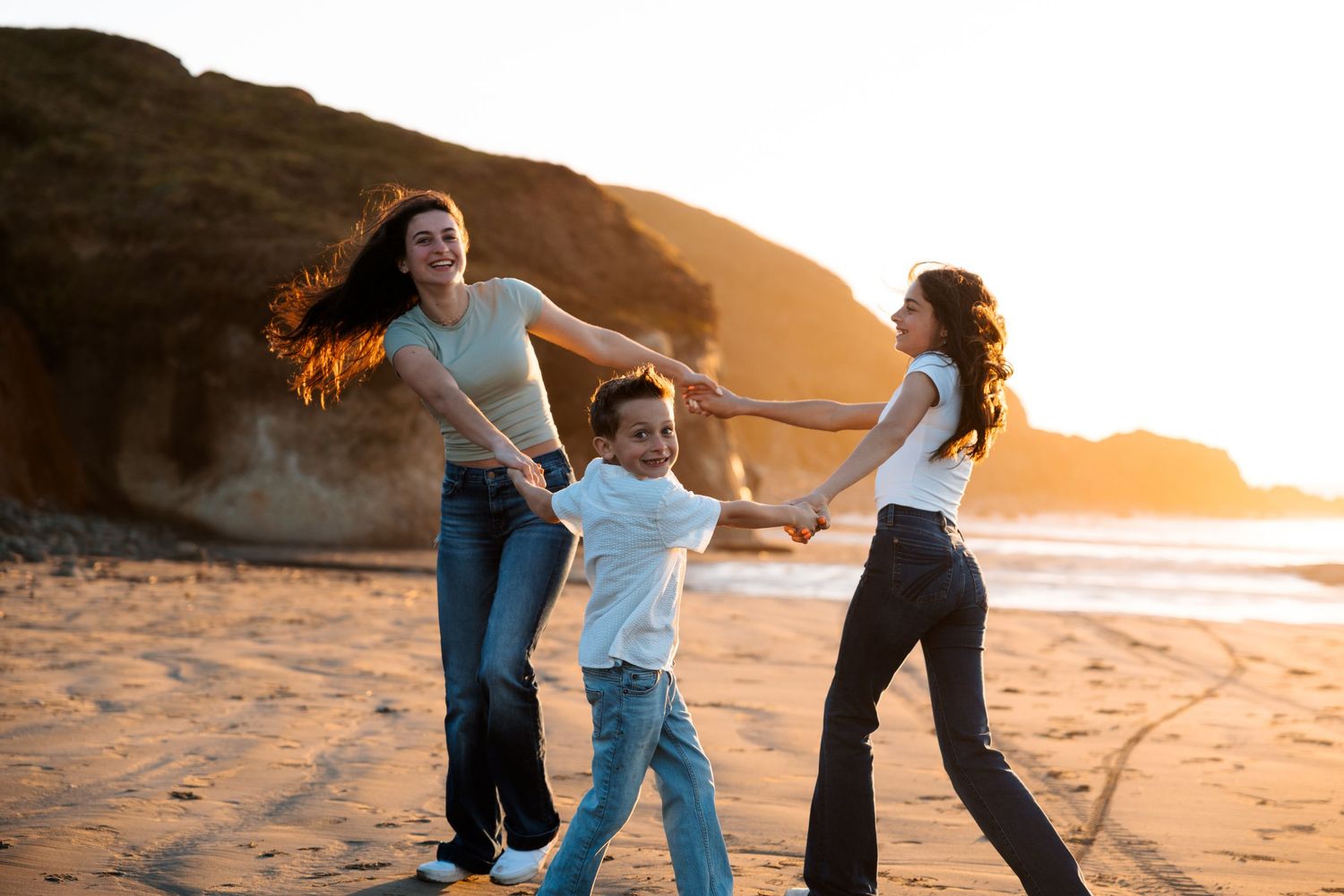 Two women and a young boy joyfully spinning together on a sandy beach at golden sunset.