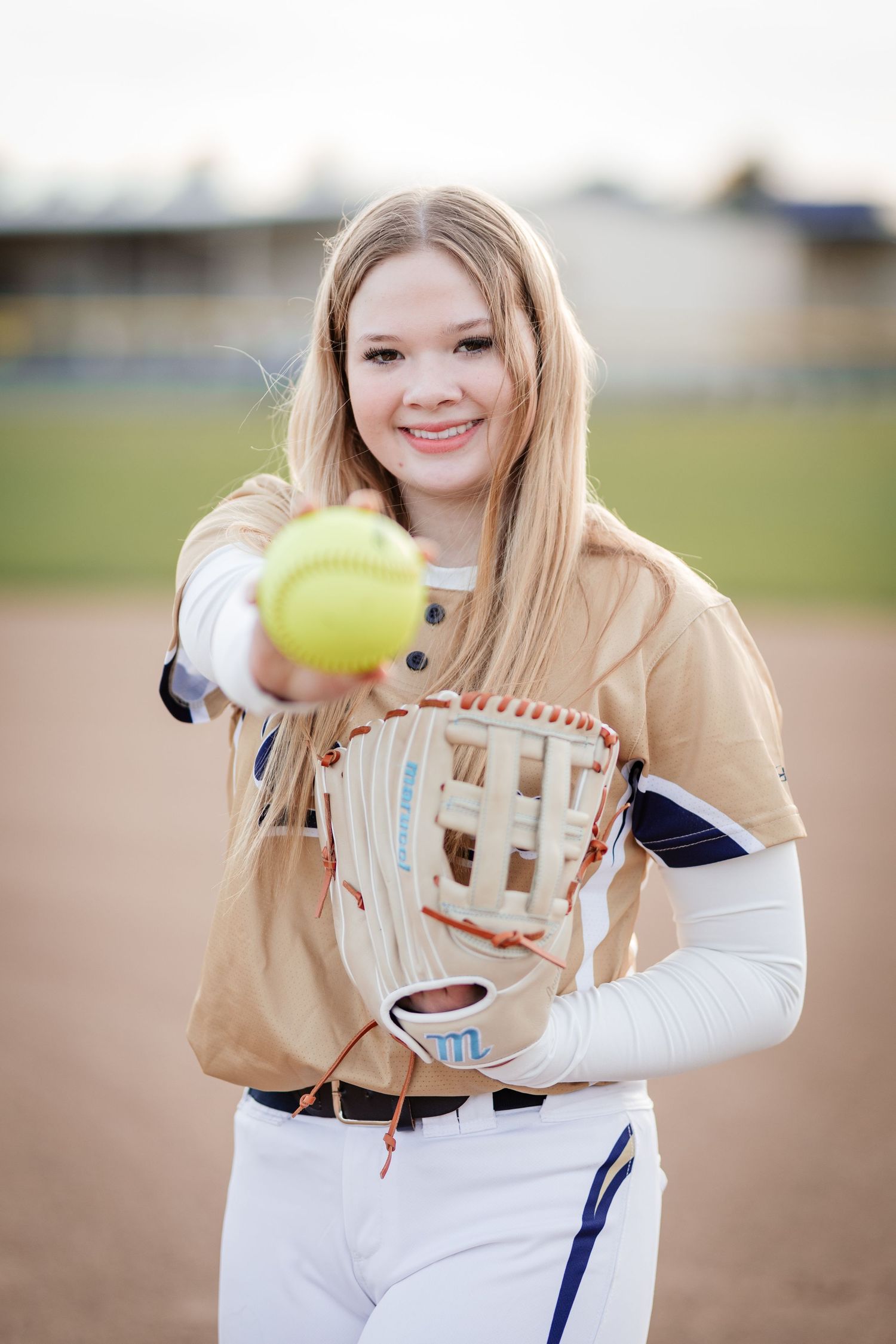 Blonde softball player extending yellow ball toward camera while holding glove on field.