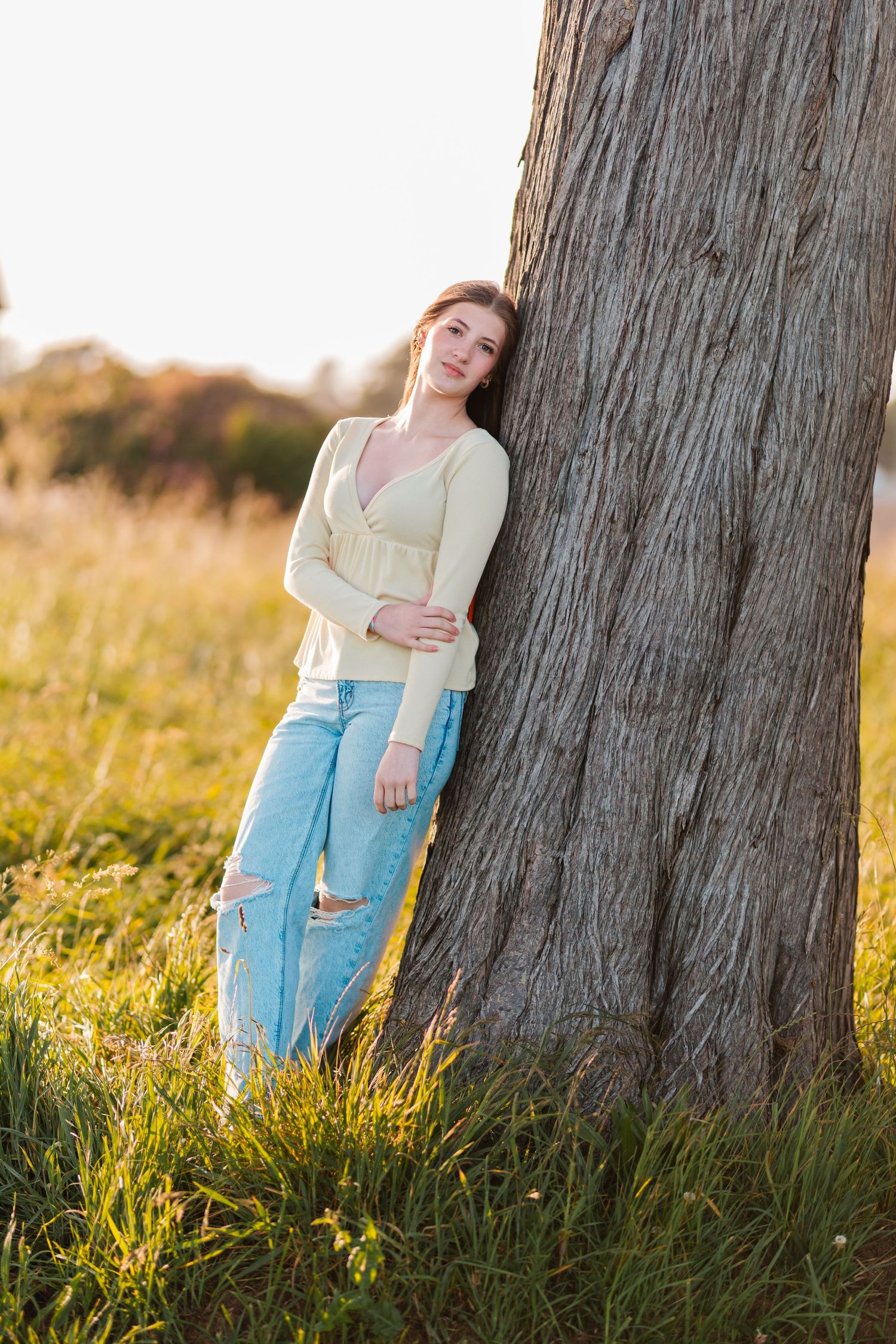 Young woman in yellow top and ripped jeans leans against large tree trunk in sunny meadow.