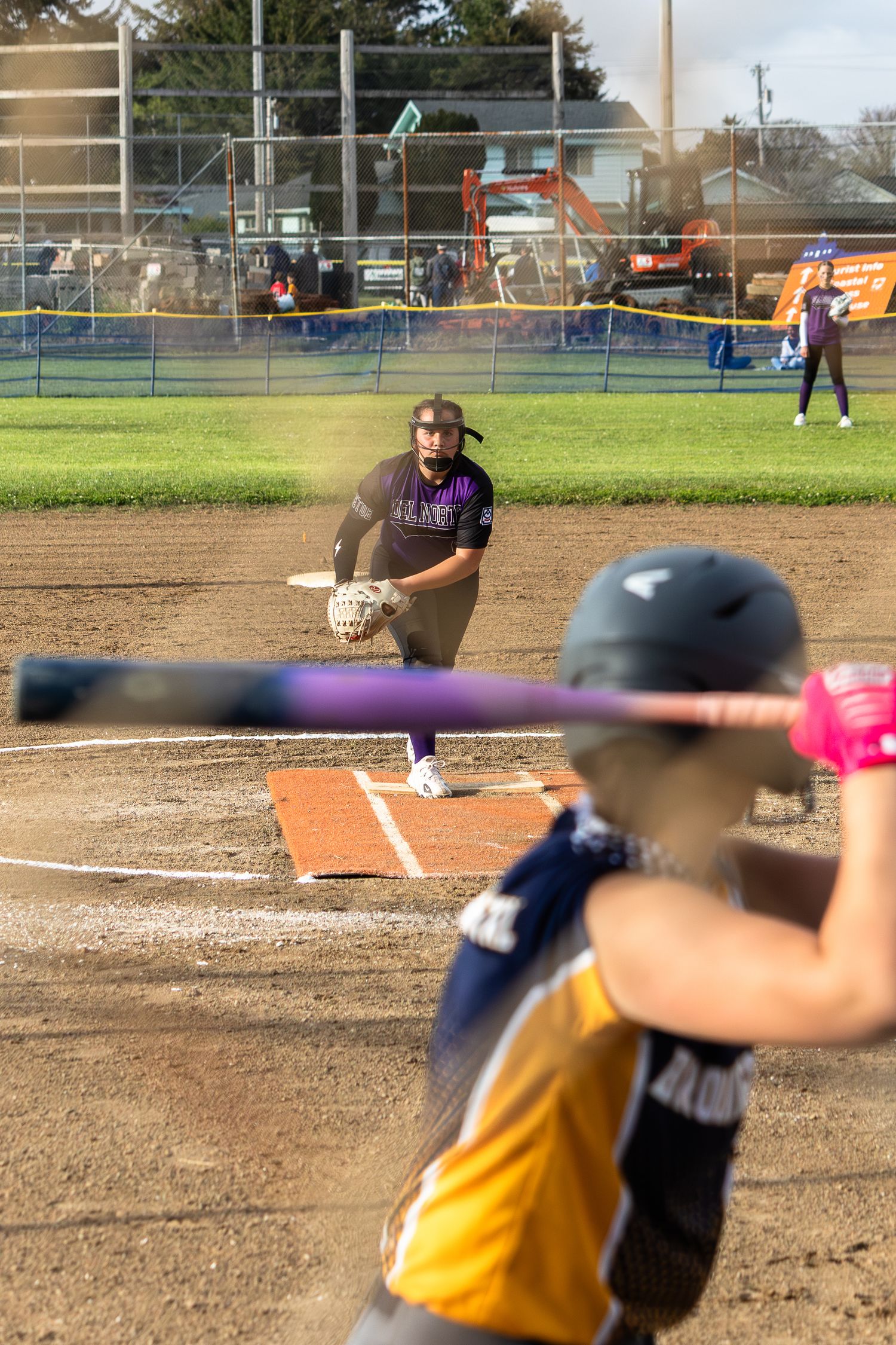 Softball pitcher in purple uniform prepares to catch as batter swings during a competitive outdoor game.