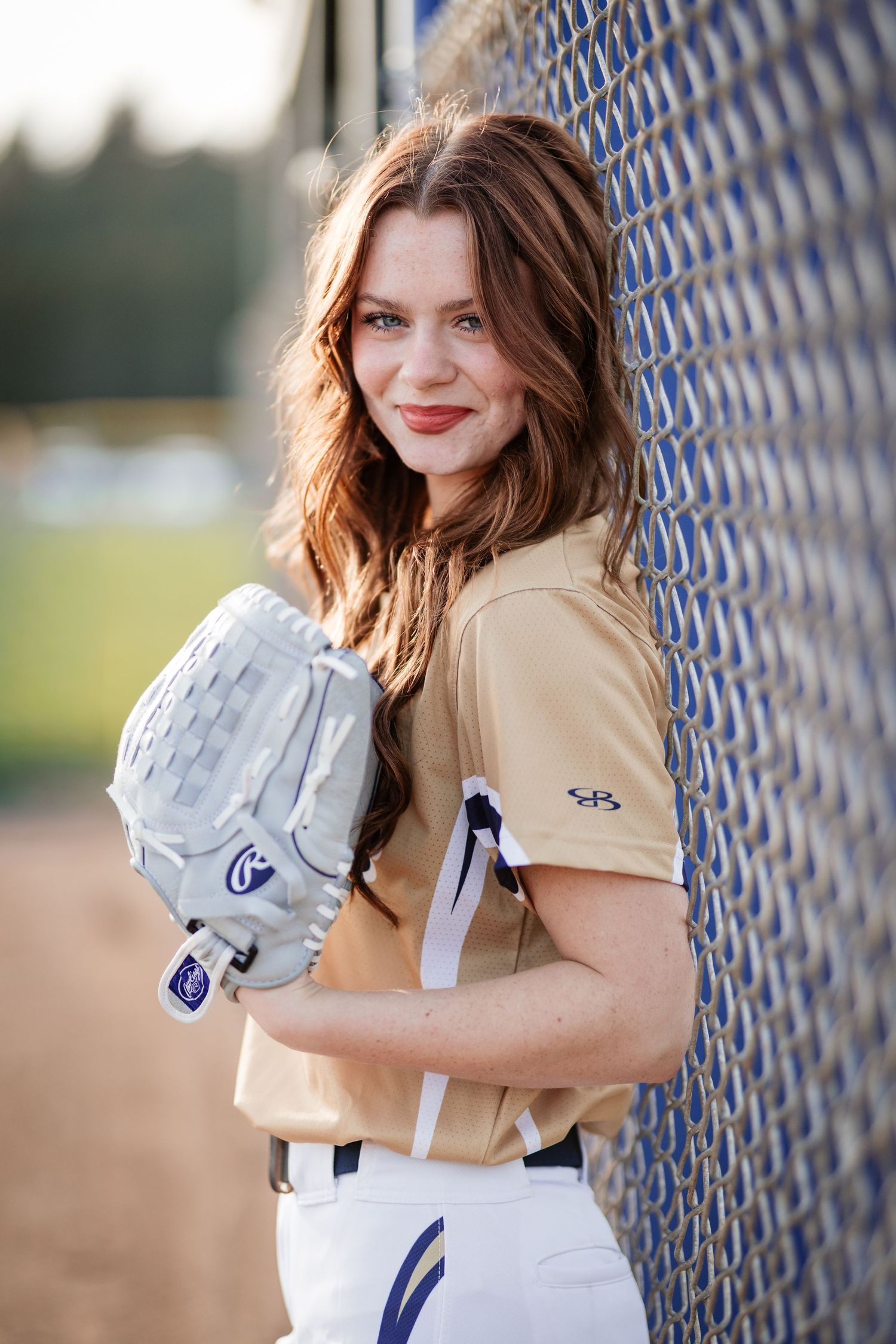 Female softball player in tan uniform leans against chain-link fence holding glove.