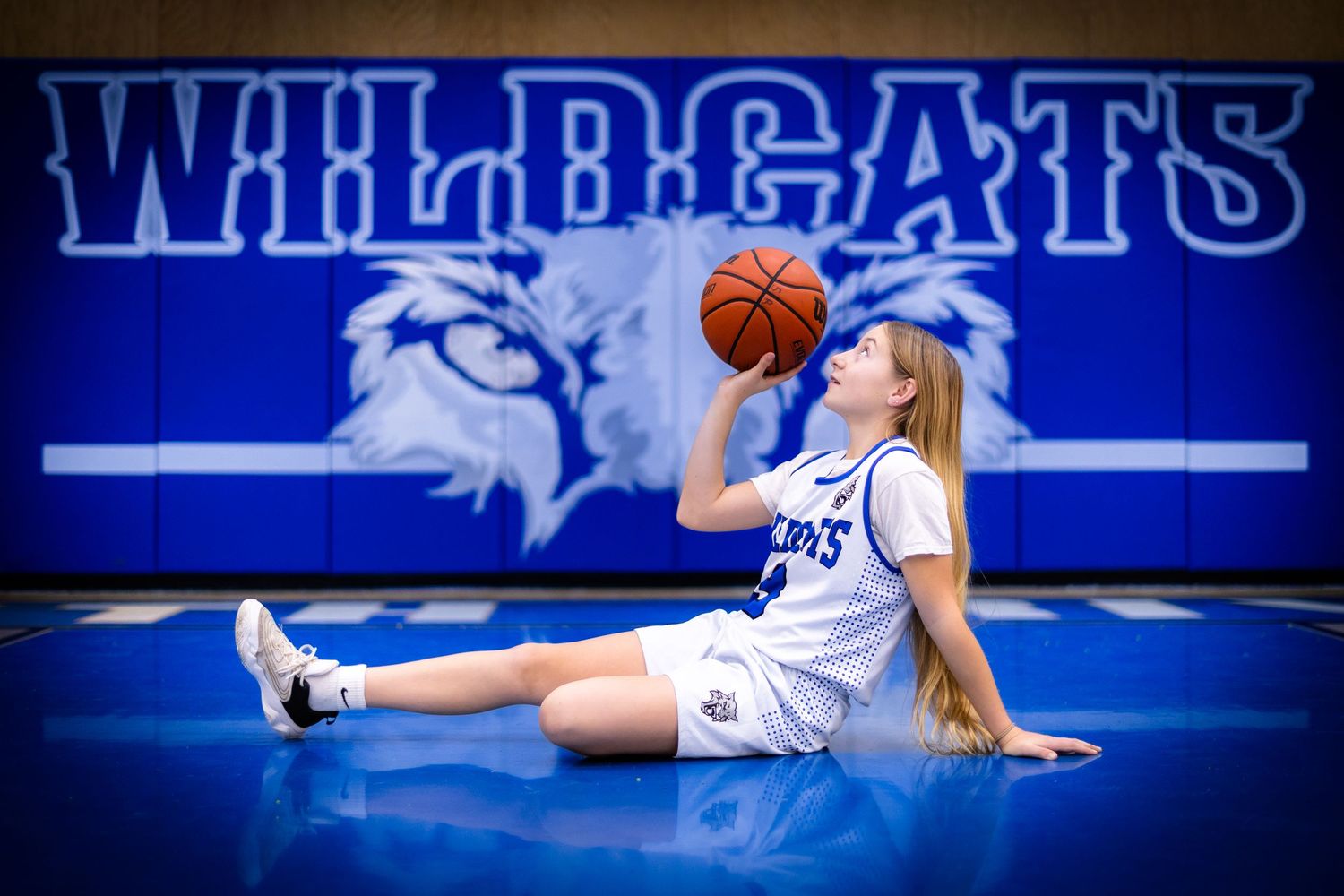 A Wildcats basketball player sits on the gym floor balancing a basketball while posing.