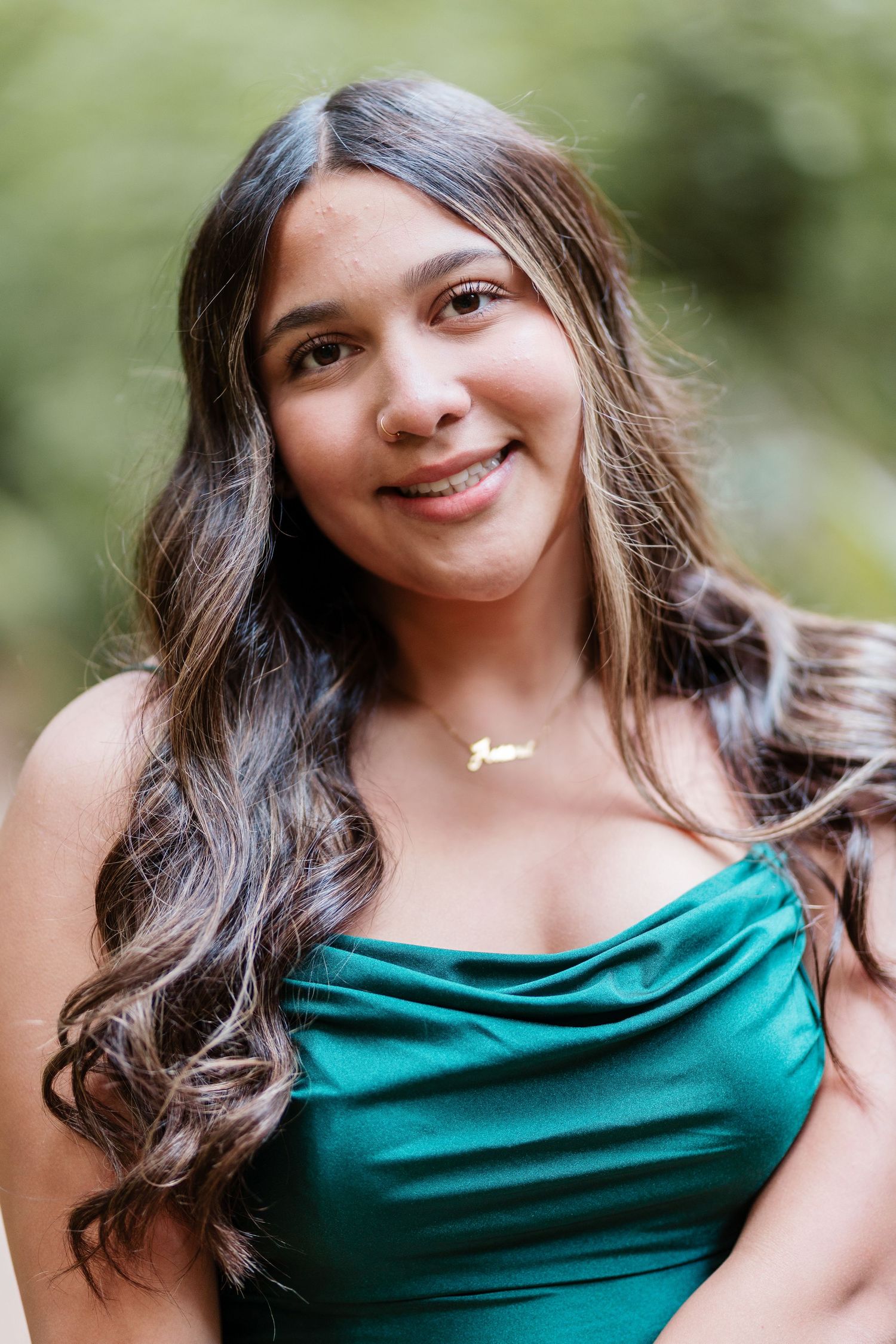 Young woman in teal strapless dress with gold necklace, smiling outdoors with lush green background.