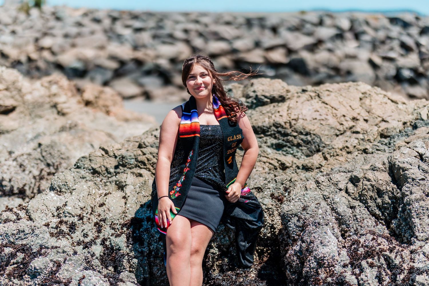 Young woman in black dress with colorful scarf sitting on rocky coastal shoreline on a sunny day.