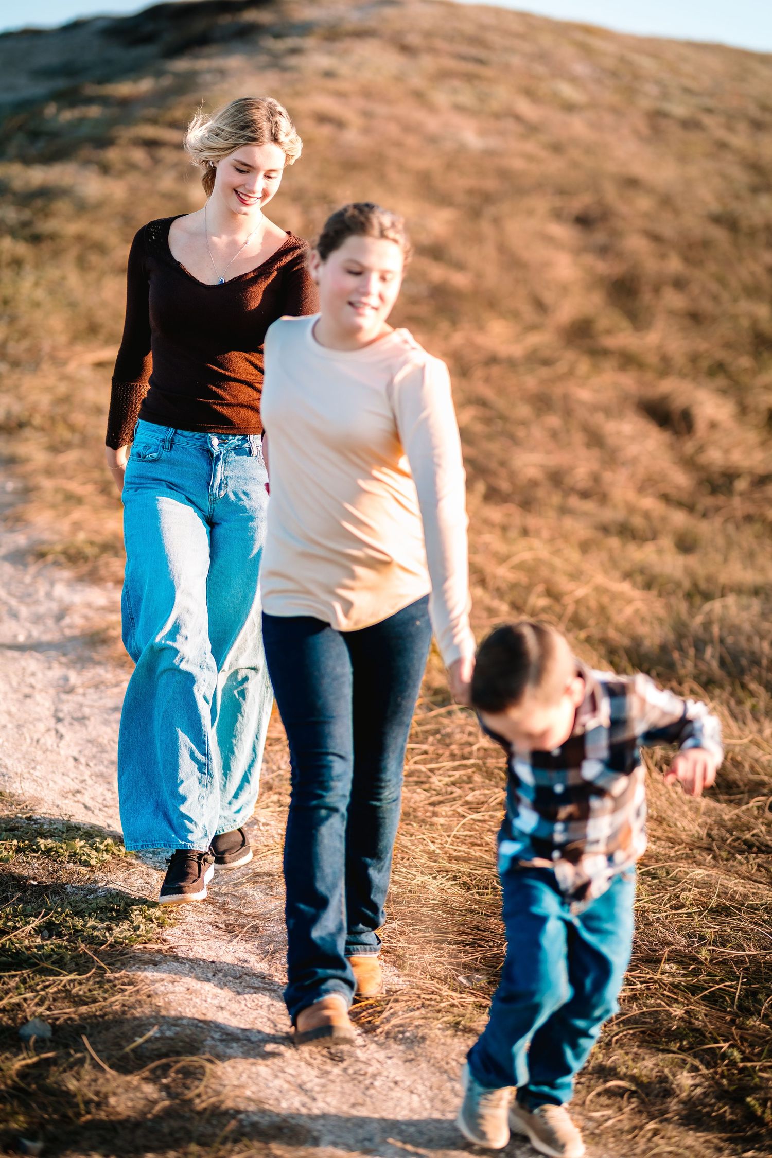 Two women and a young boy walk along a sunny dirt path on a hillside, smiling and enjoying the outdoors.