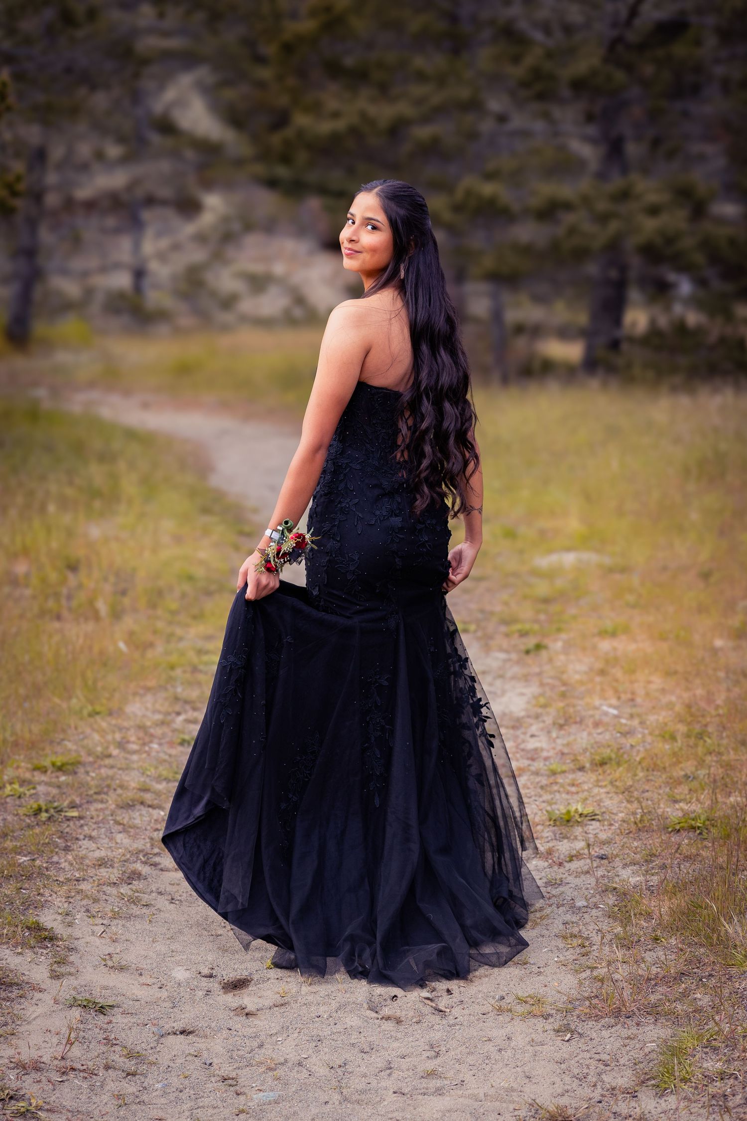 Young woman in black mermaid prom dress with colorful wrist corsage posing from behind on a scenic dirt path.